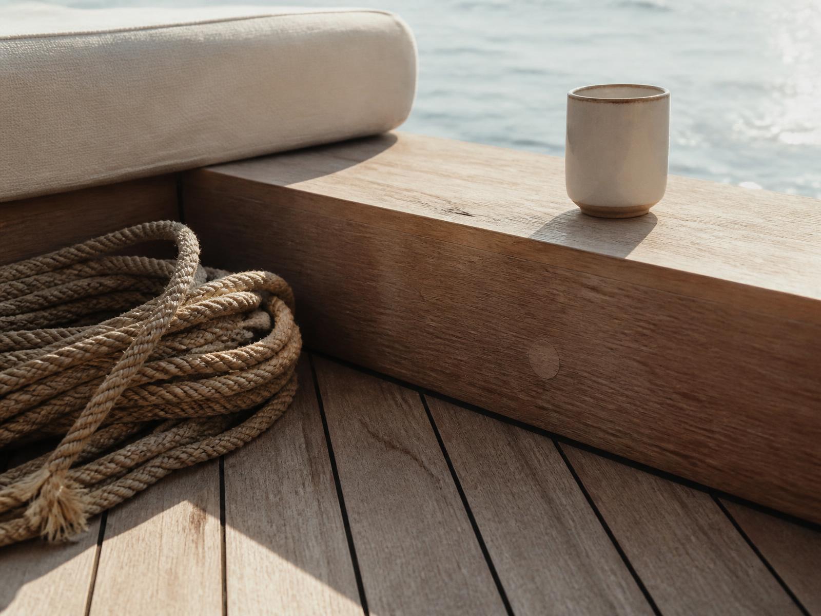 Teak deck, a coiled rope and a single ceramic cup on a pale timber bench in morning sea light