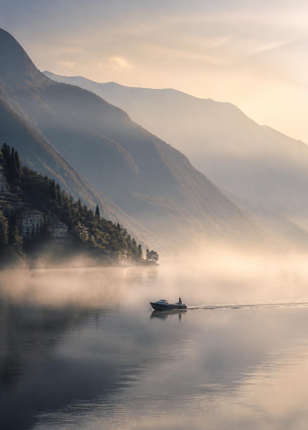 Morning mist rising off Lake Como with a small boat crossing still silver water and mountains fading in layers