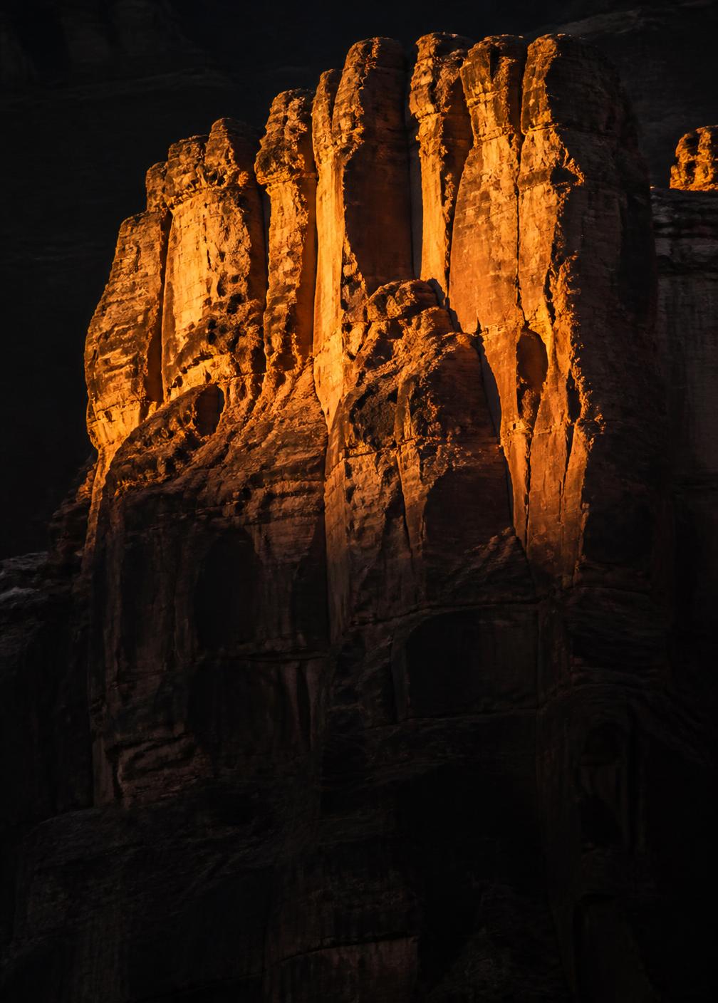 First light striking the top of a towering sandstone cliff face while the base remains in deep shadow
