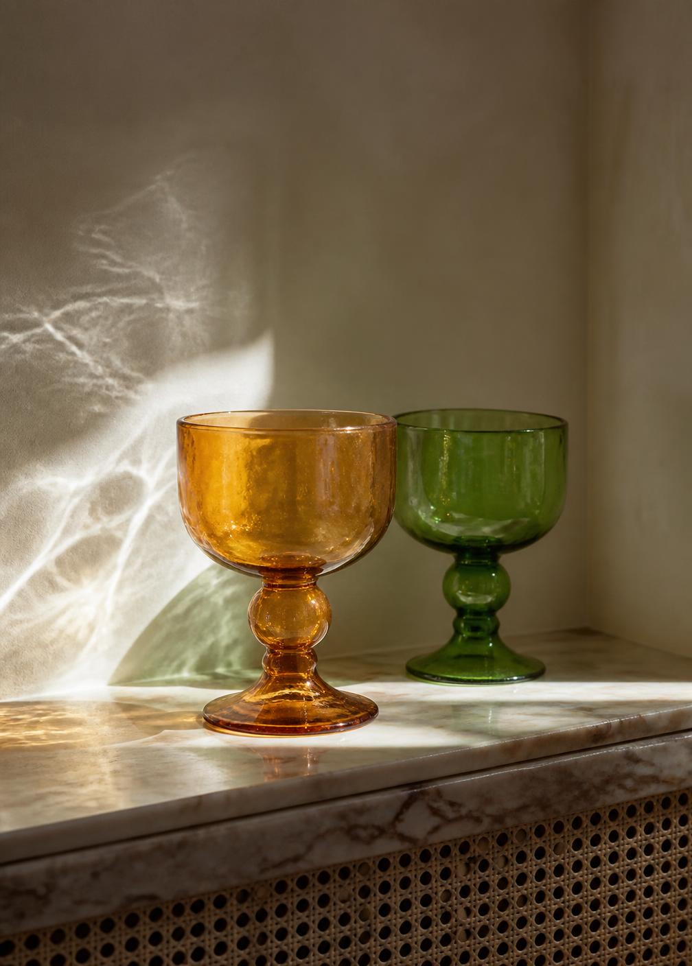 Two hand-blown Murano glass goblets in amber and green on a marble ledge with water reflections on the wall behind