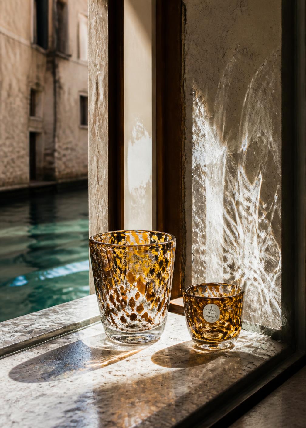 Two amber Murano glasses on a marble ledge beside a canal window with water reflections on the wall