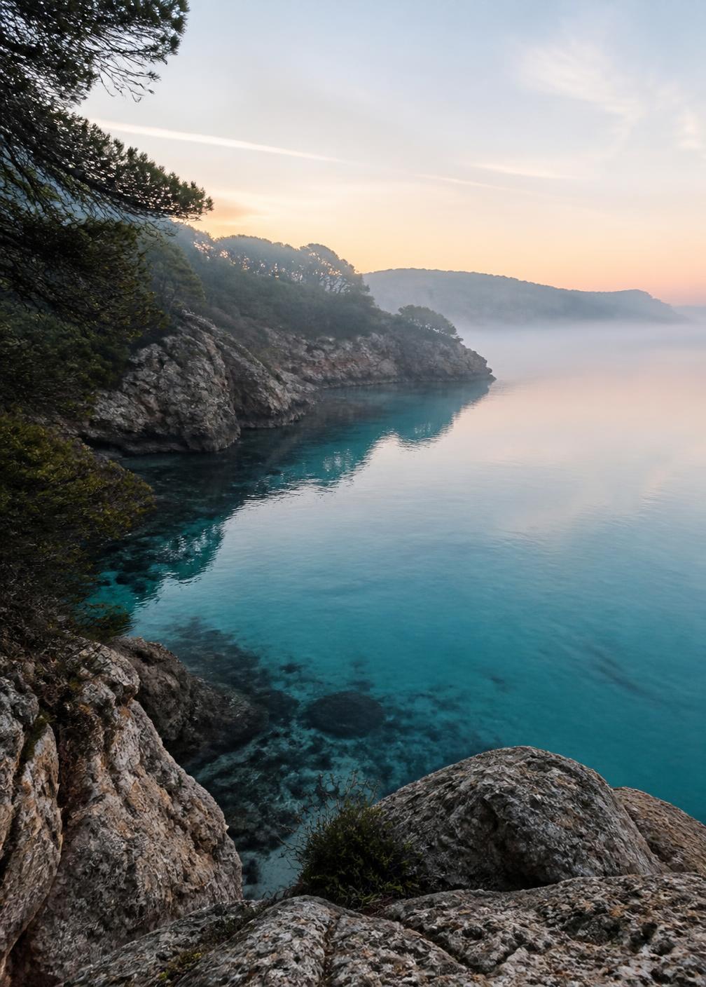Still turquoise cove water at dawn in northern Ibiza seen from limestone rocks with faint mist over the sea