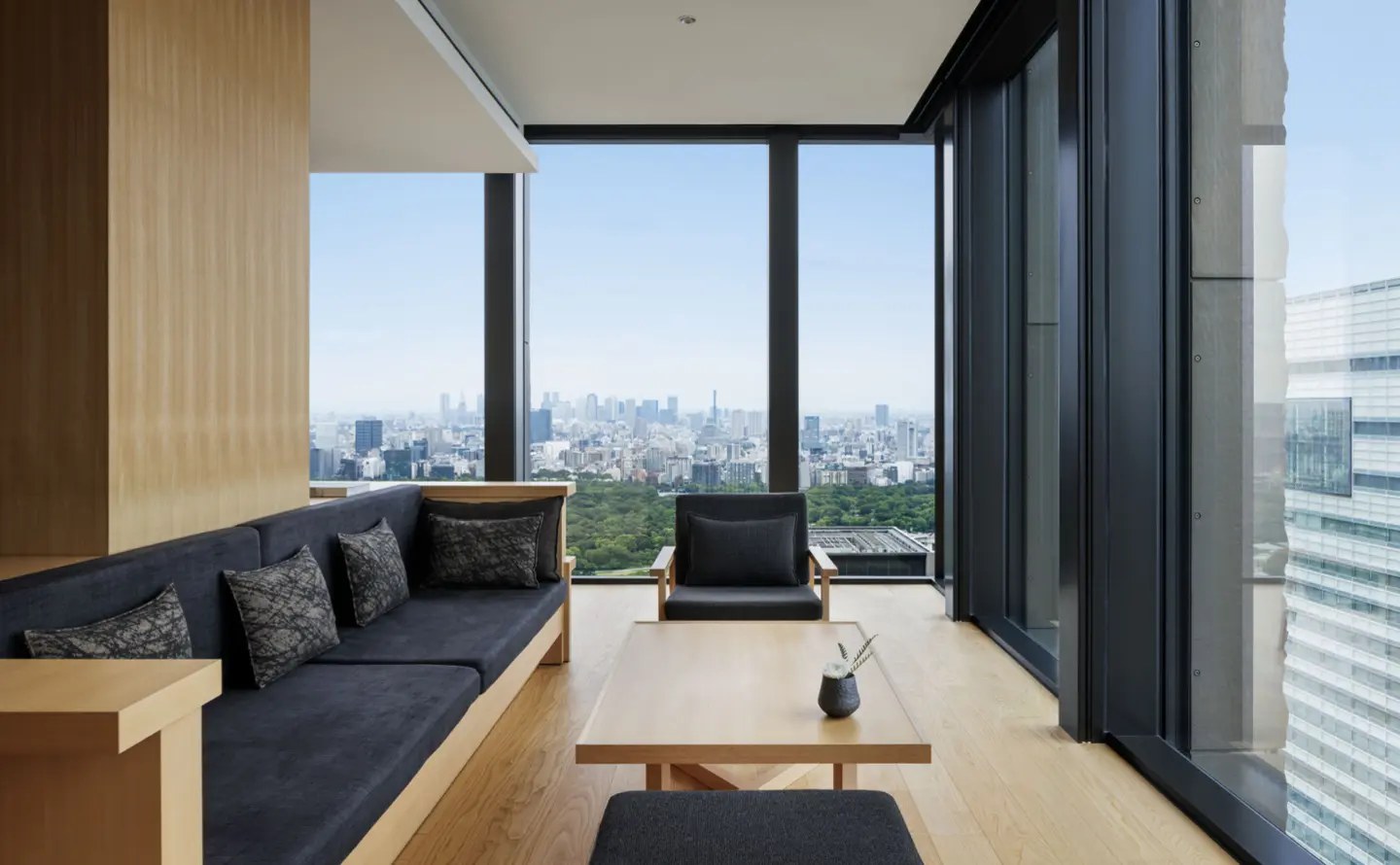 Suiten-Lounge mit heller Holzvertäfelung, dunklem Sofa und Eckfenster über den Baumkronen Tokios