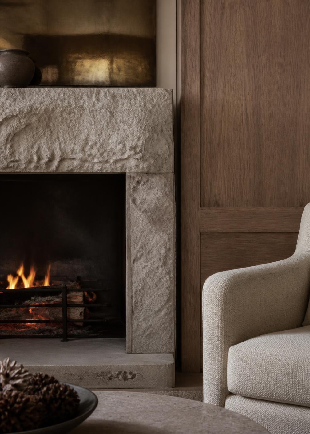 Rough-hewn stone fireplace with a low flame beside oak panelling and a linen armchair