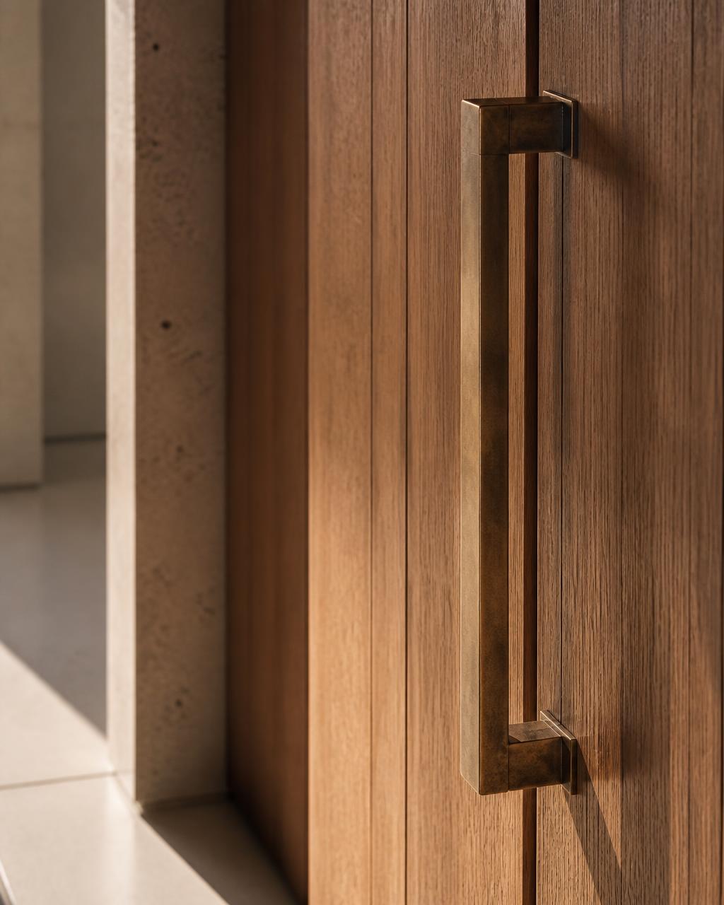Close detail of a hand-finished bronze door pull against warm oak panelling with pale stone flooring in soft morning daylight