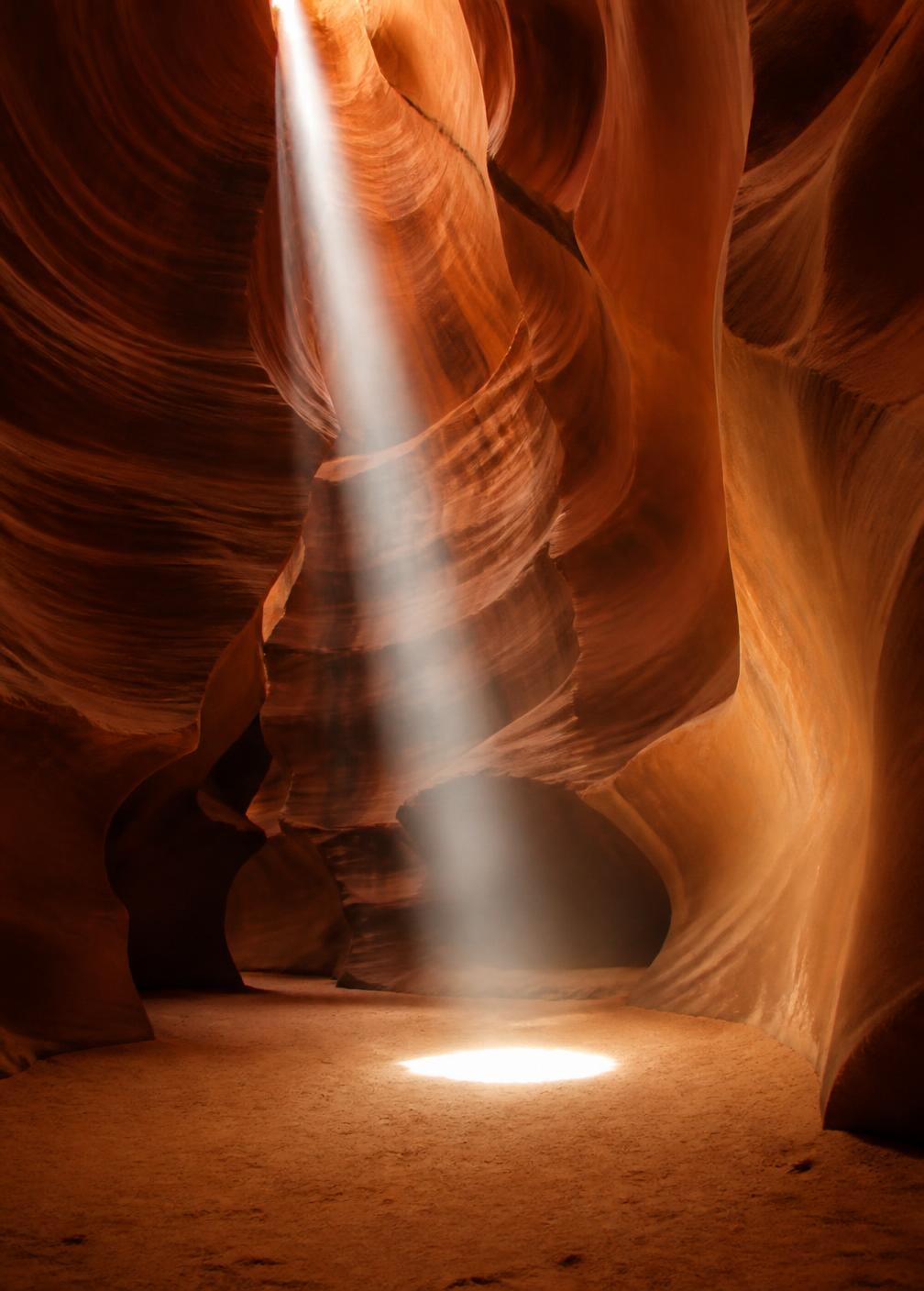 Interior of a narrow slot canyon with sculpted ochre walls and a single shaft of light falling to the sand floor