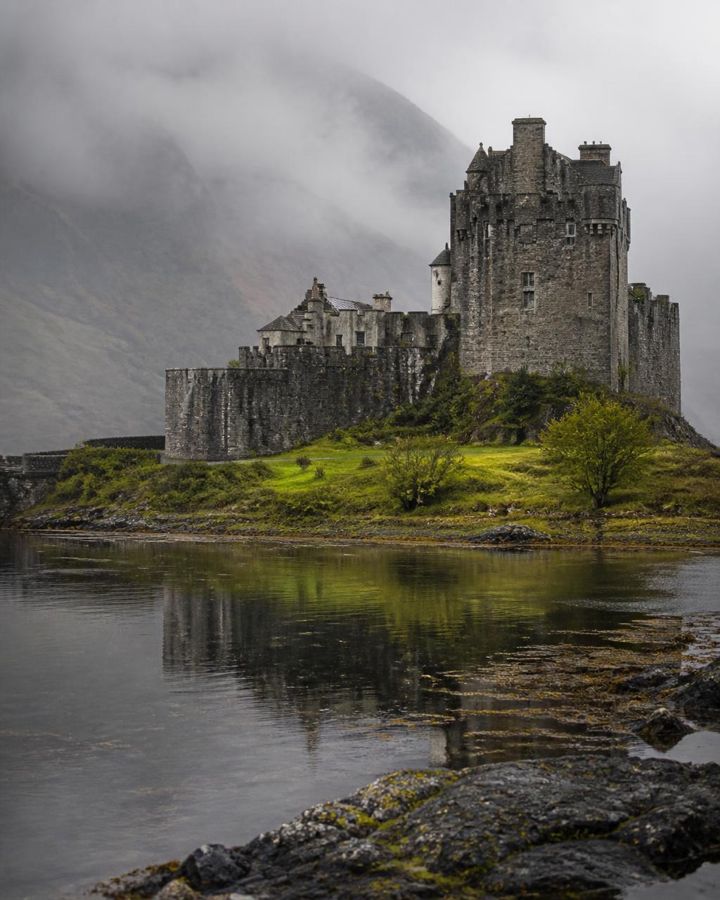 Ein graues schottisches Steinschloss erhebt sich bei Morgendämmerung über stillem Loch-Wasser, tiefe Wolken über den Hügeln dahinter