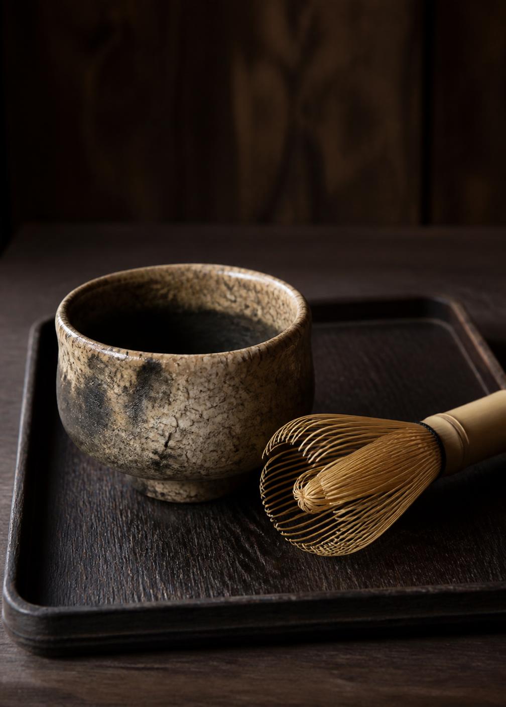Raku tea bowl and bamboo whisk on a dark timber tray in low side light