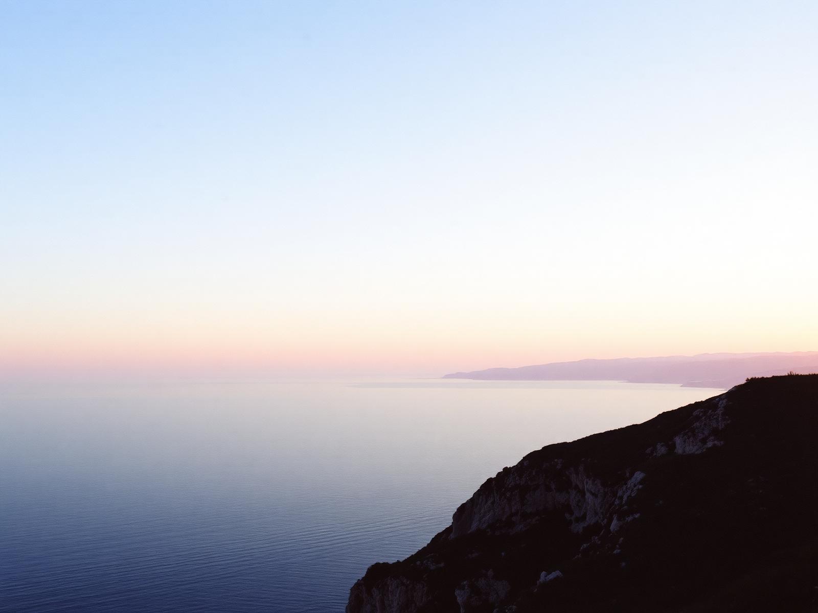 Calm Mediterranean sea and headland at dusk under a pale cream and rose sky
