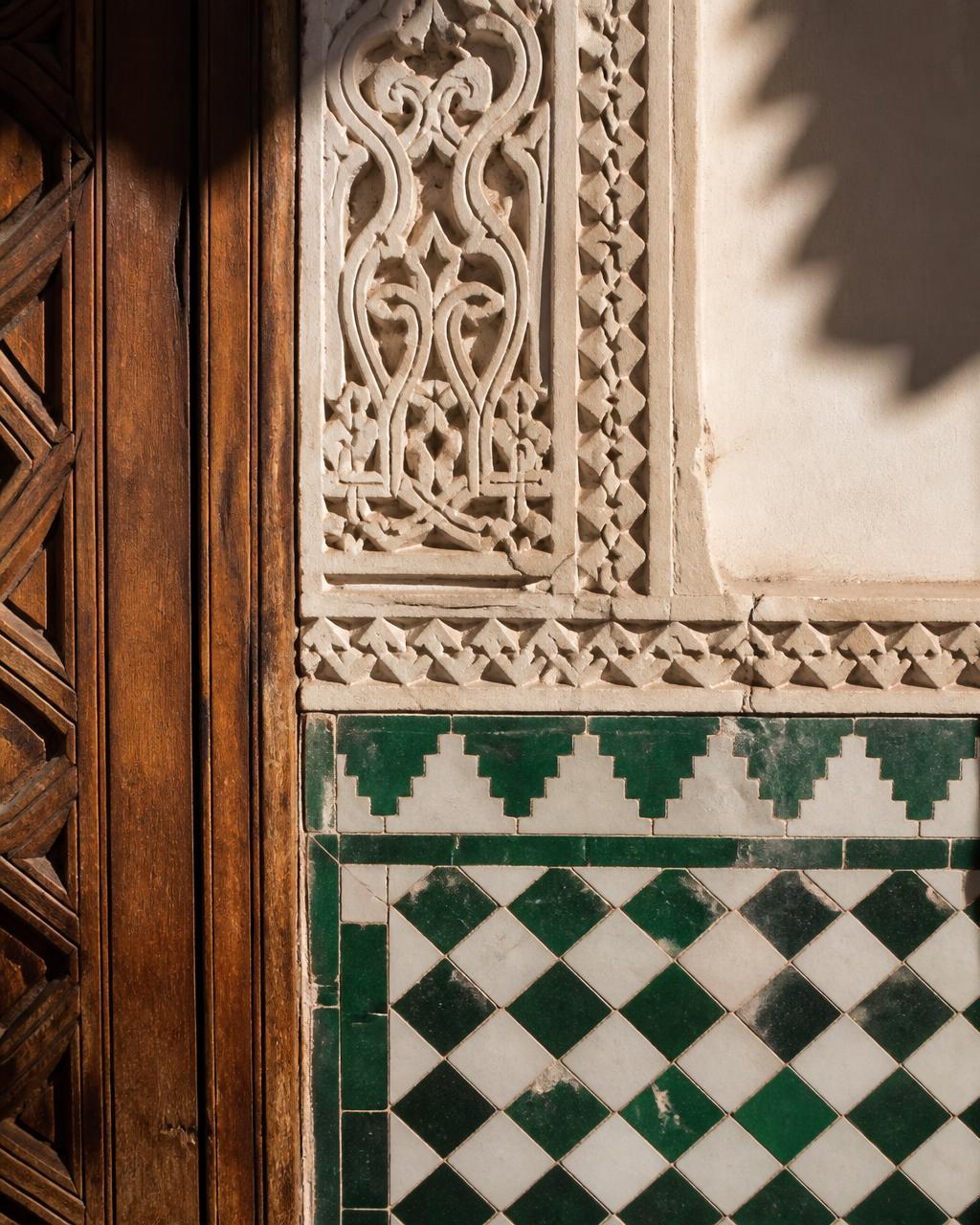 Close detail of green and white hand-cut zellige tilework meeting carved stucco and old cedar wood in Morocco