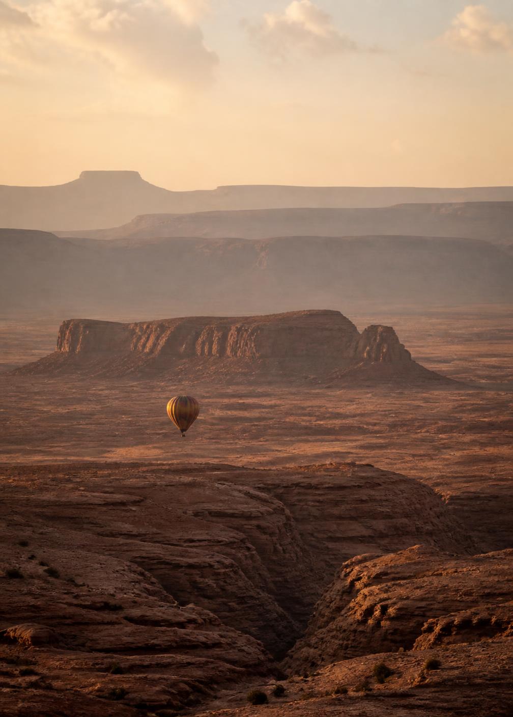 A single hot-air balloon drifting low over vast red desert mesas in sunrise haze
