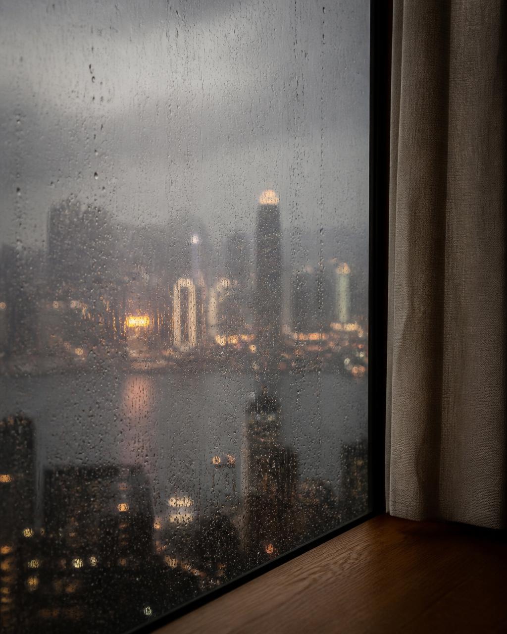 Rain running down a floor-to-ceiling window high above Hong Kong with the skyline dissolved into soft grey and amber light
