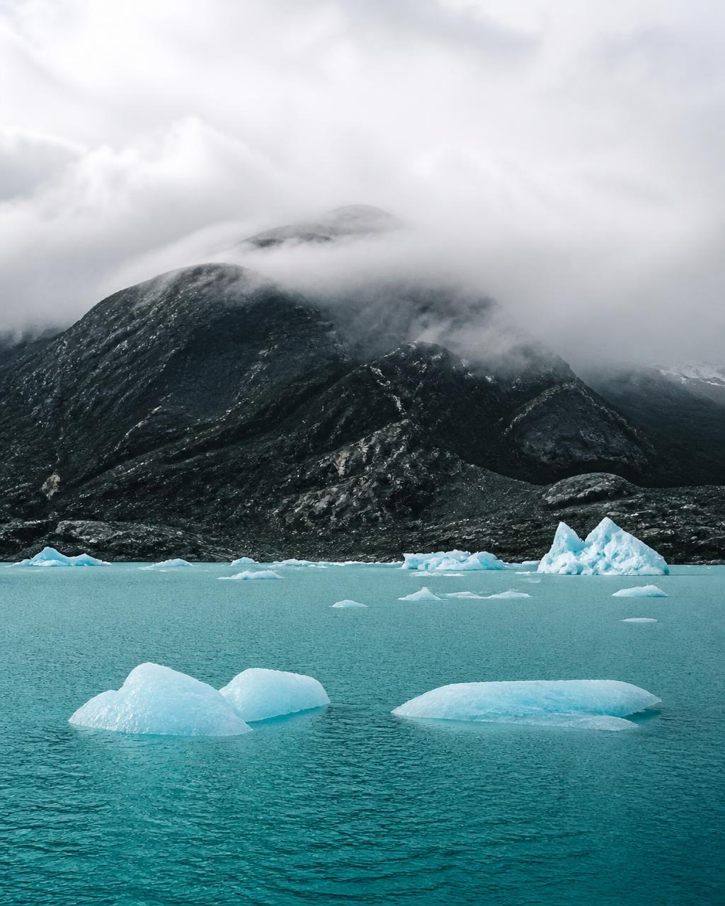 A turquoise glacial lake in Patagonia with floating pale blue ice and low cloud pouring over a dark ridge
