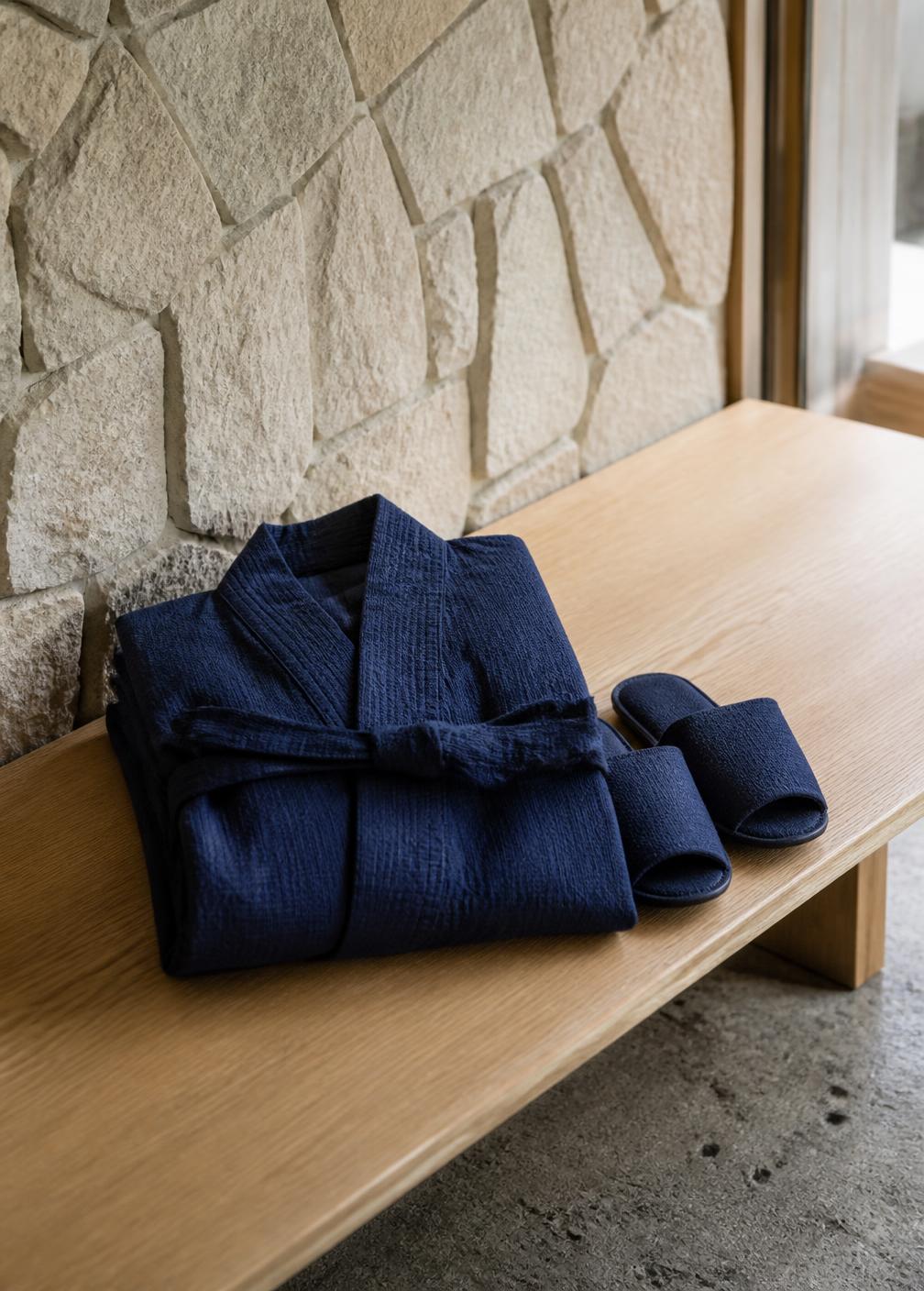 Folded indigo cotton robe and slippers on a pale wood bench beside a rough stone wall