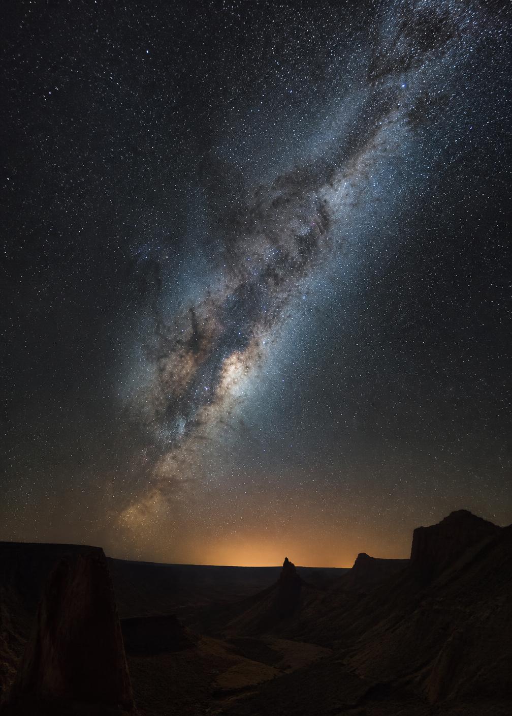 The Milky Way arching over the dark silhouette of a desert canyon under a star-filled sky