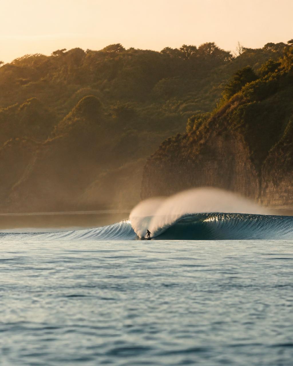 A long clean left-hand wave peeling across an empty reef bay in Sumba at golden hour with green cliffs behind