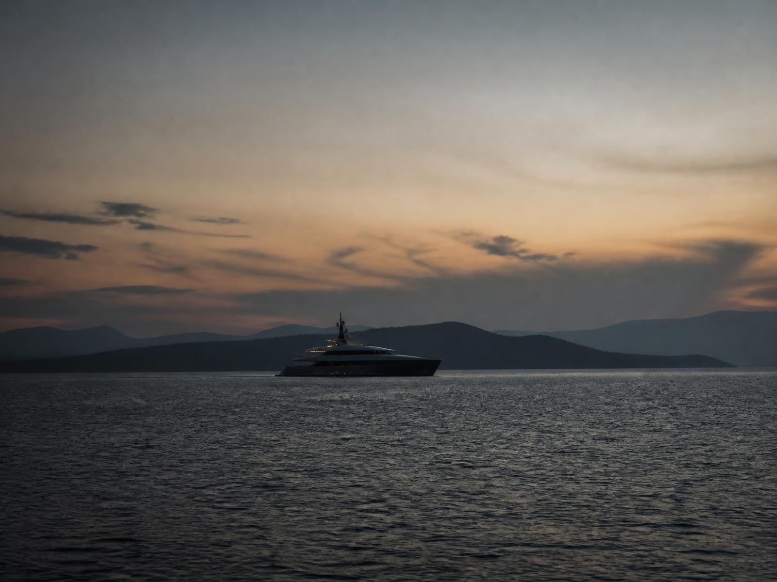 Dark yacht seen small against a vast dusk sea and a distant island silhouette