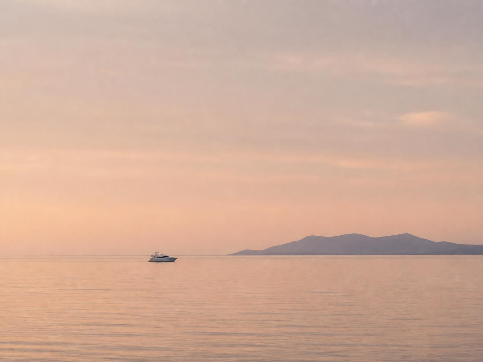 A small white yacht seen distant against a vast pale dusk sea and a faint island silhouette