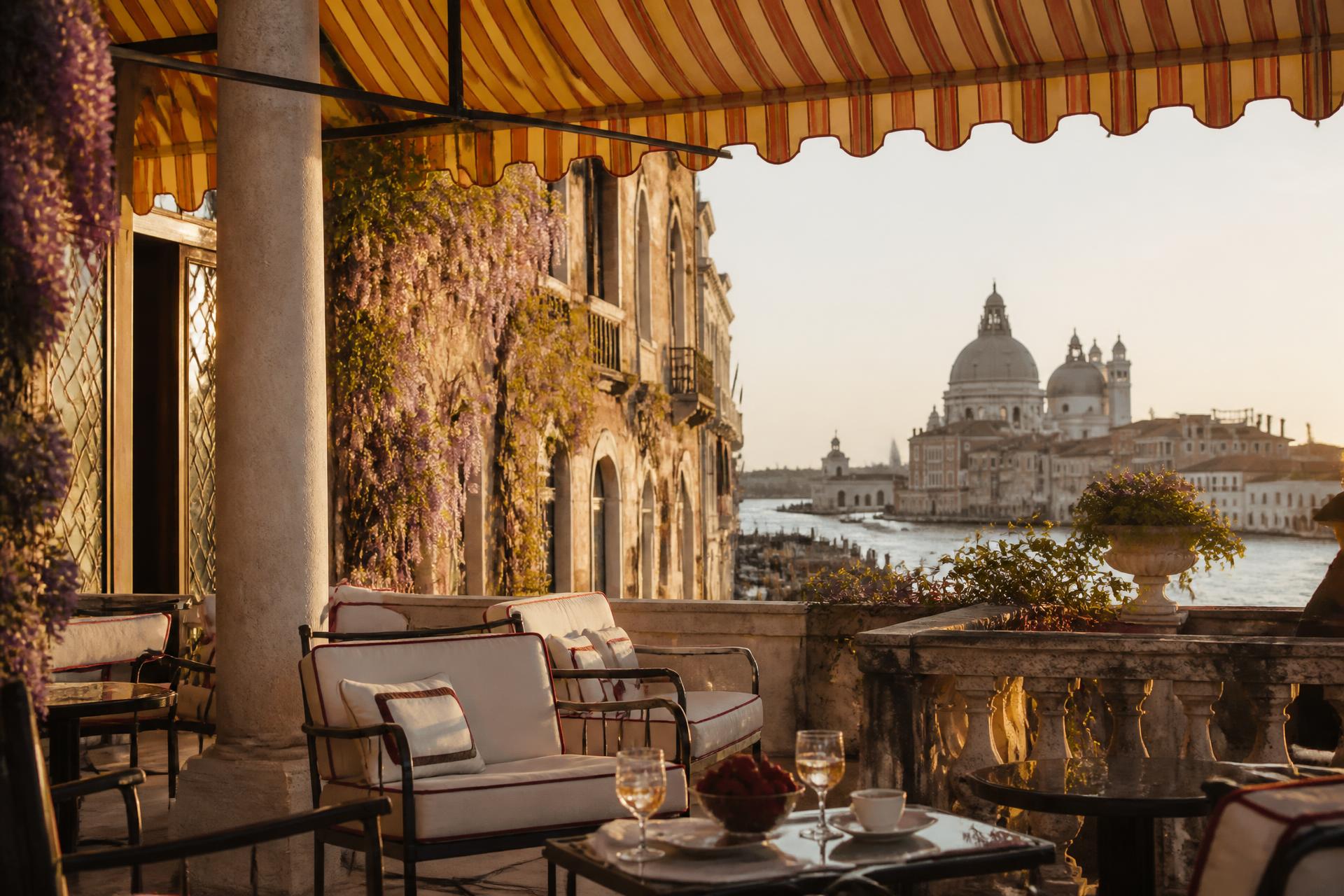Venetian palazzo terrace under a striped awning with wisteria and a view across the lagoon to a domed basilica at golden hour