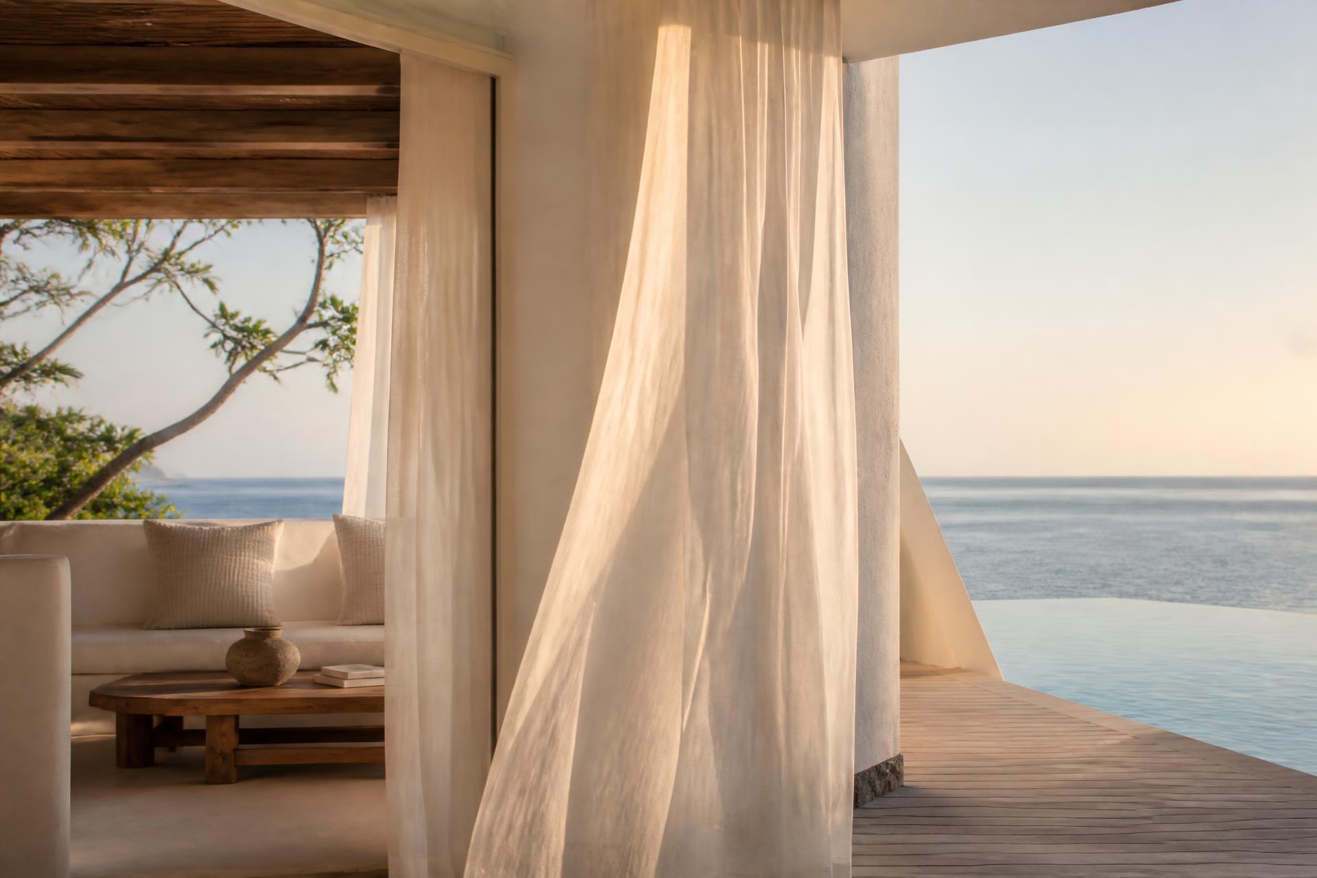 White island pavilion opening onto a calm sea, sheer linen curtains drifting over a pale teak deck at golden hour