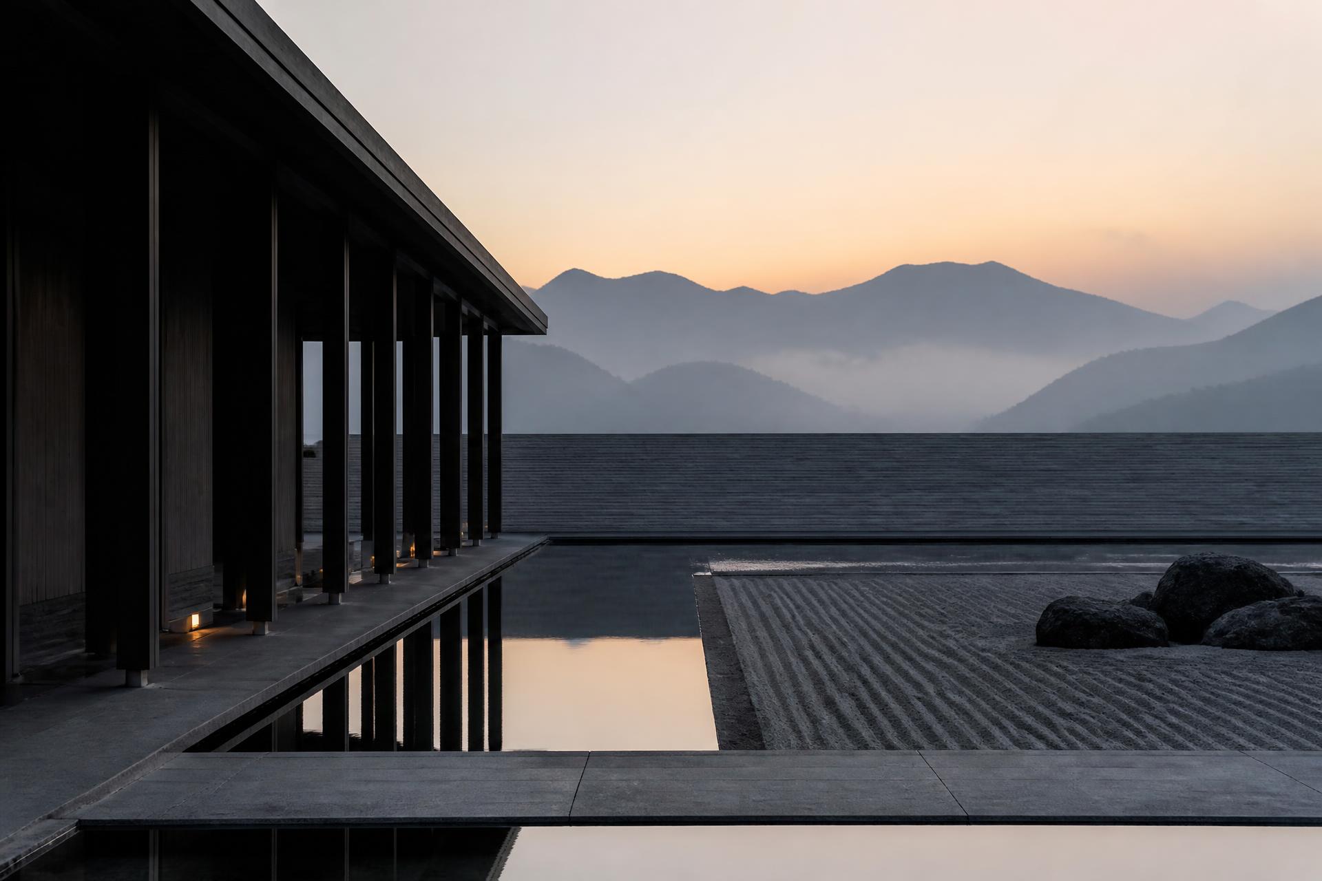 Dark timber colonnade beside a still reflecting pool and raked stone court, mountains dissolving in dawn mist