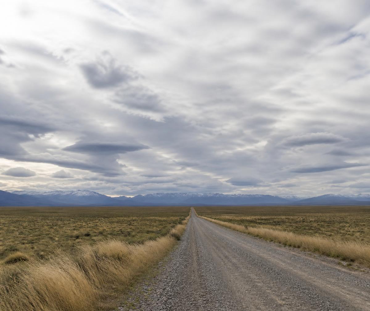 Endless Patagonian steppe under an enormous cloud-filled sky with a single dirt track running to the horizon and distant blue mountains