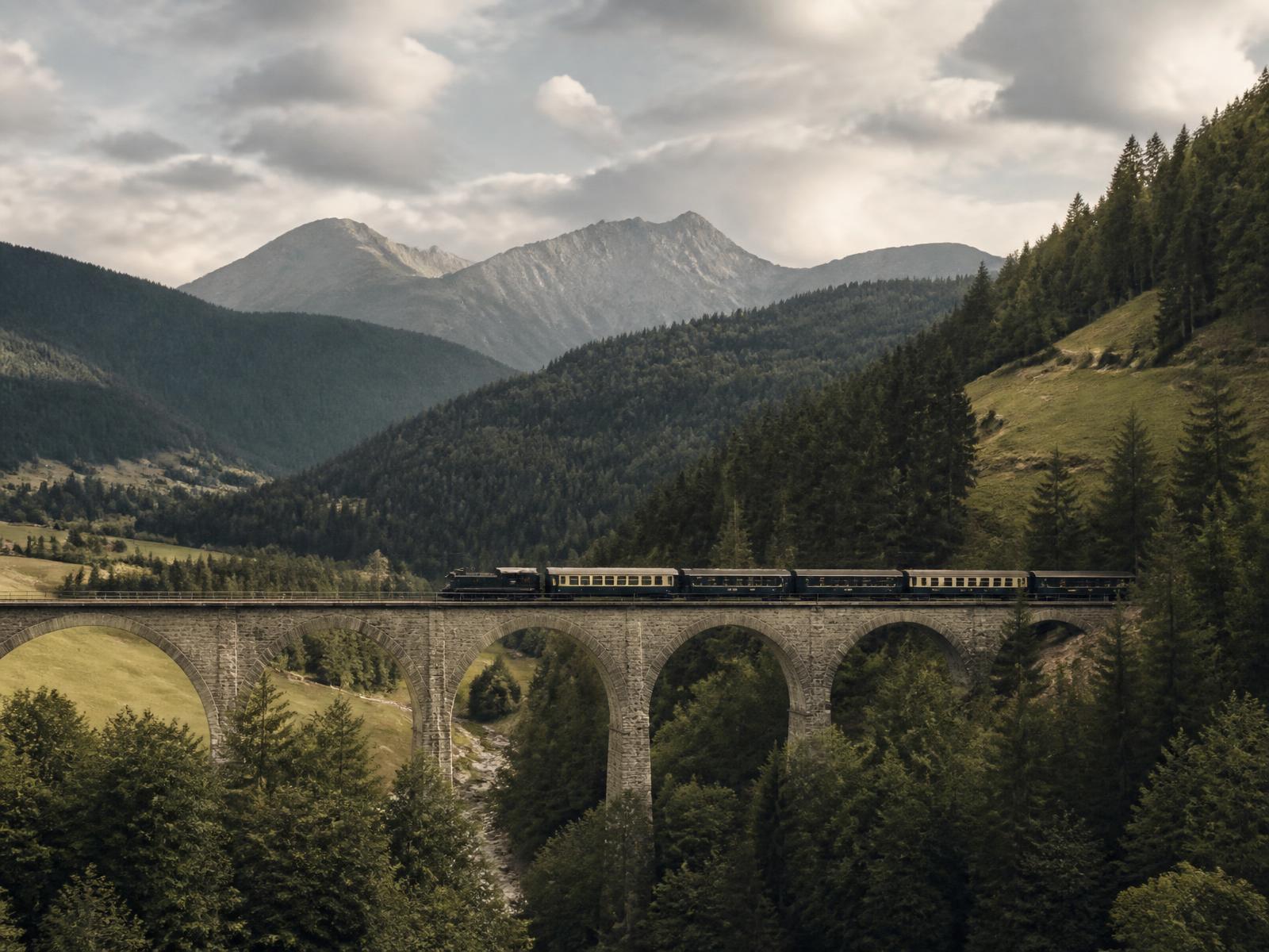 A vintage blue-and-gold luxury train crossing a stone viaduct through green European mountains in soft afternoon light