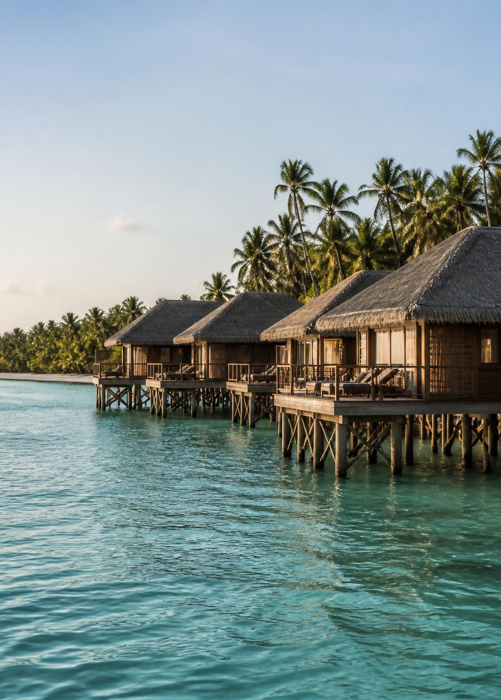 A row of thatched overwater villas on timber stilts above a calm turquoise Maldivian lagoon in morning light