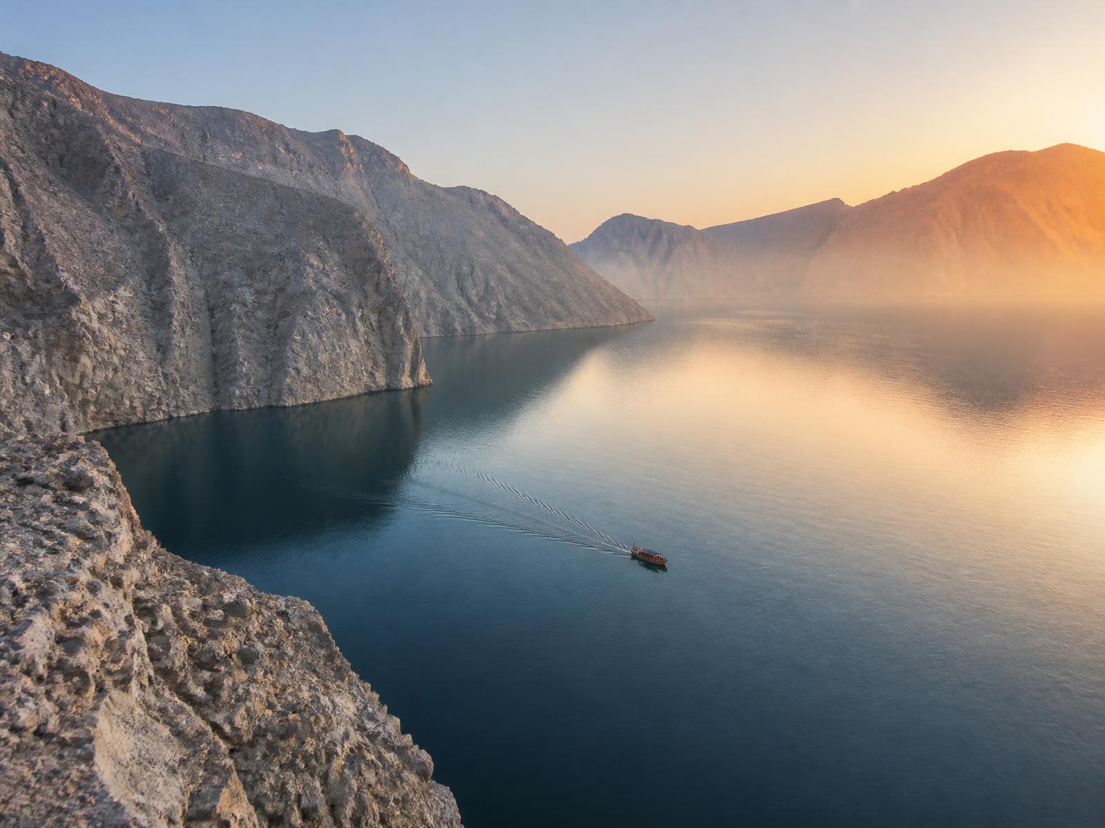 Sheer bare rock walls of the Musandam fjords falling into deep still water at dawn with a small wooden boat far below