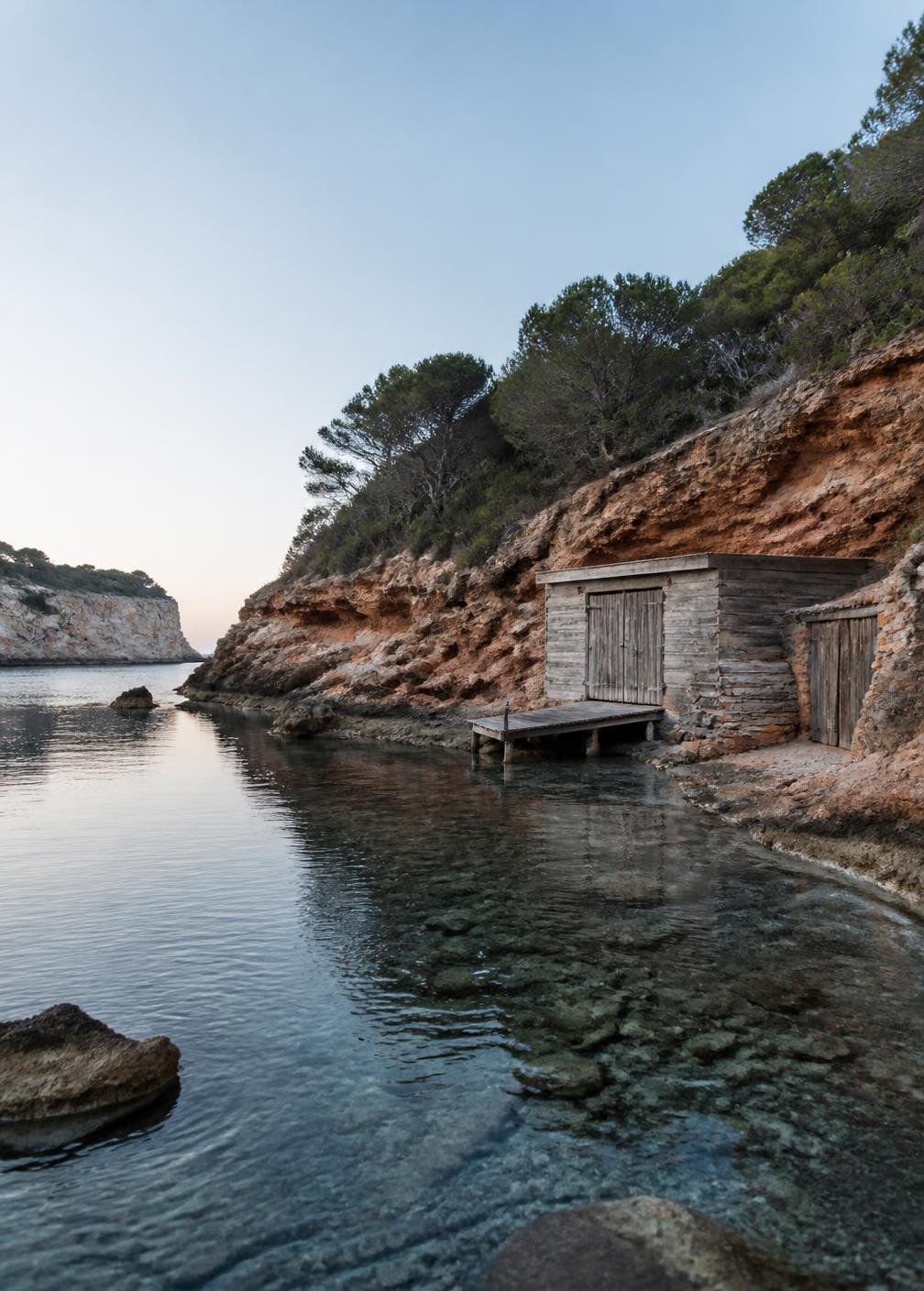 Hidden cove in northern Ibiza with red rocks, clear water and a small wooden fishing hut in early morning light