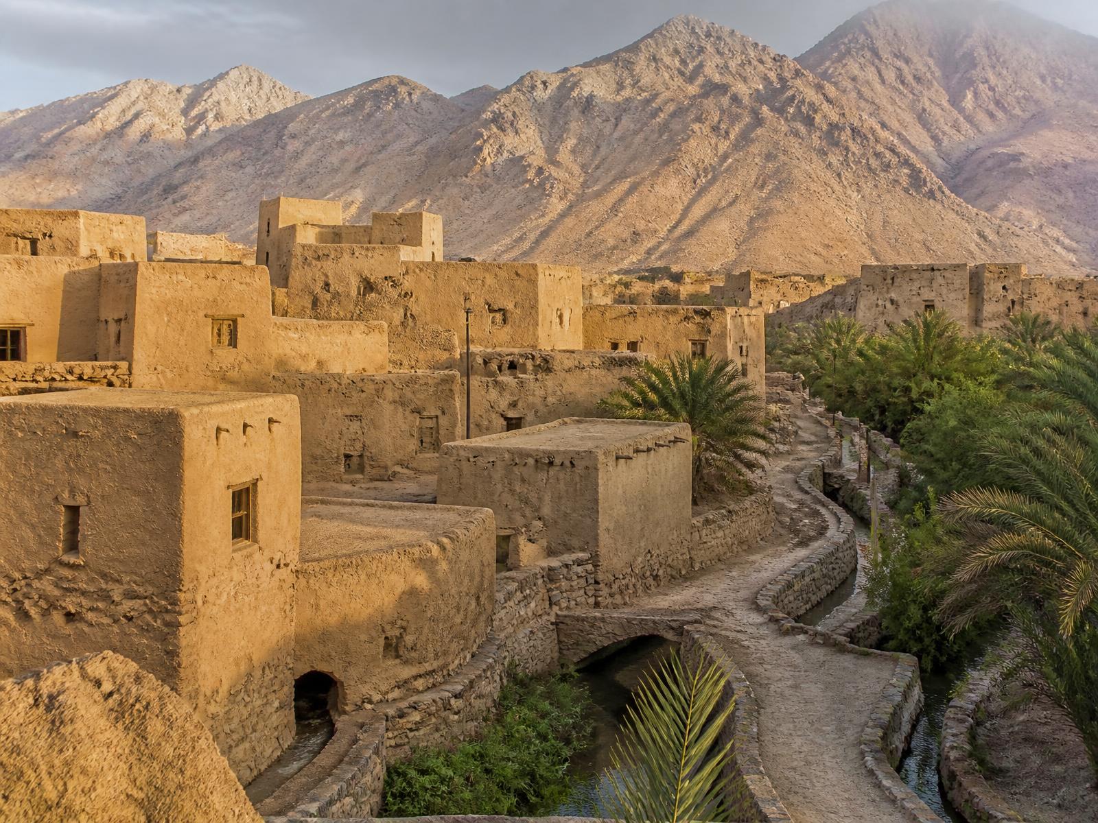An old mud-brick and stone Omani mountain village at golden hour with flat roofs, a falaj water channel and date palms