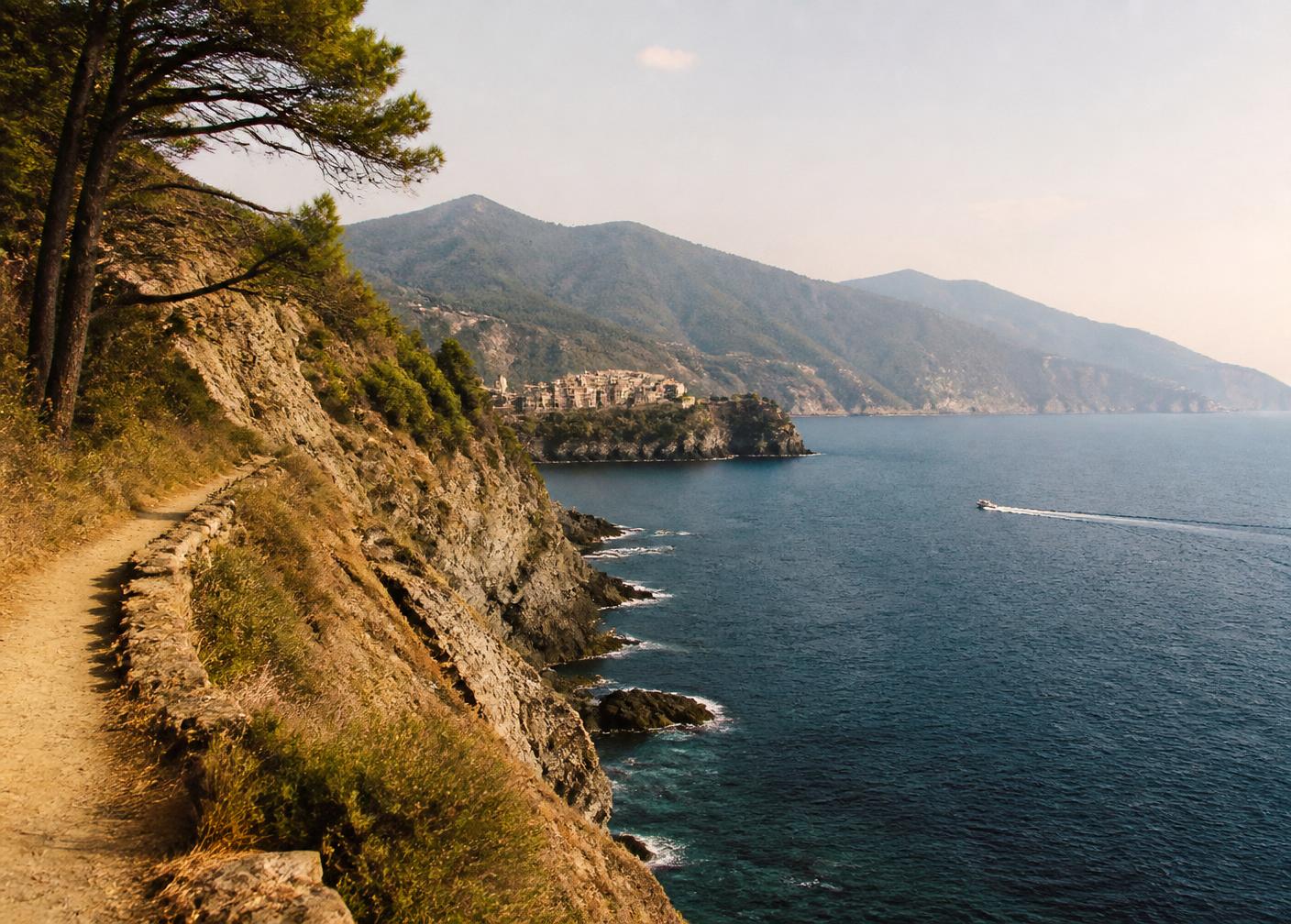 Ligurian coastline seen from a pine-shaded coastal footpath with cliffs, a distant pastel village and a small boat wake