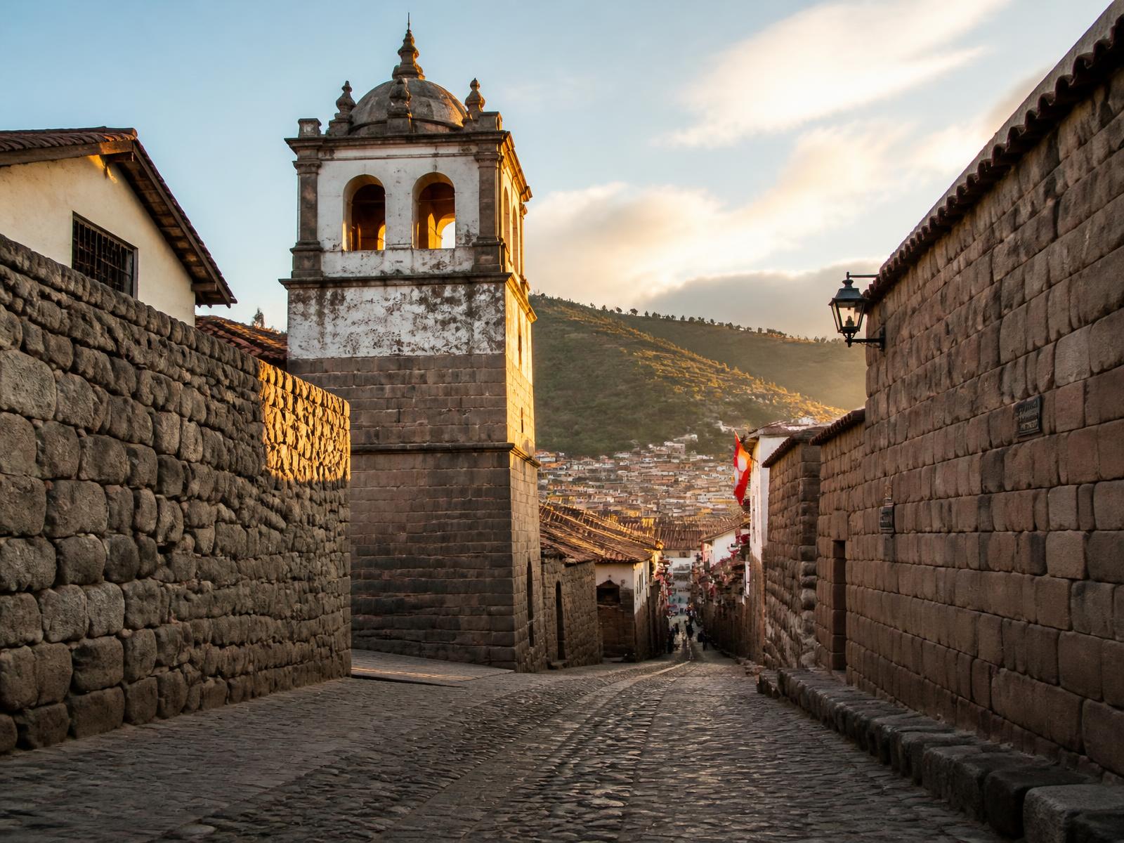 Eine gepflasterte Straße in Cusco zur goldenen Stunde mit Inka-Mauerwerk, einem kolonialen Glockenturm und Andenbergen im Hintergrund