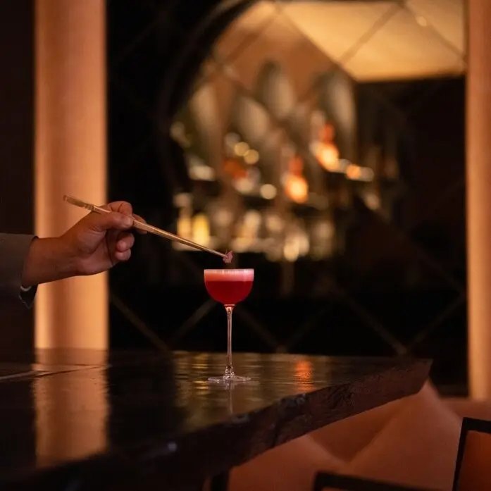 Ein Cocktail wird an der Bar des Arva in Amanjena vollendet, dahinter das gedämpfte Licht der Flaschenregale