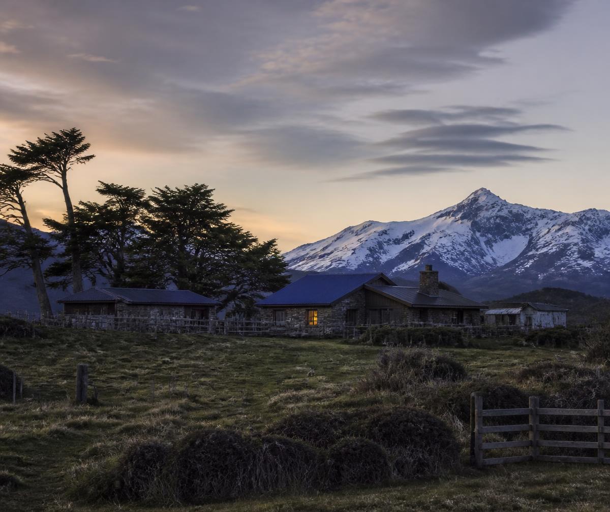 A remote Patagonian estancia at dusk with low stone and timber buildings, wind-shaped trees and snow mountains behind