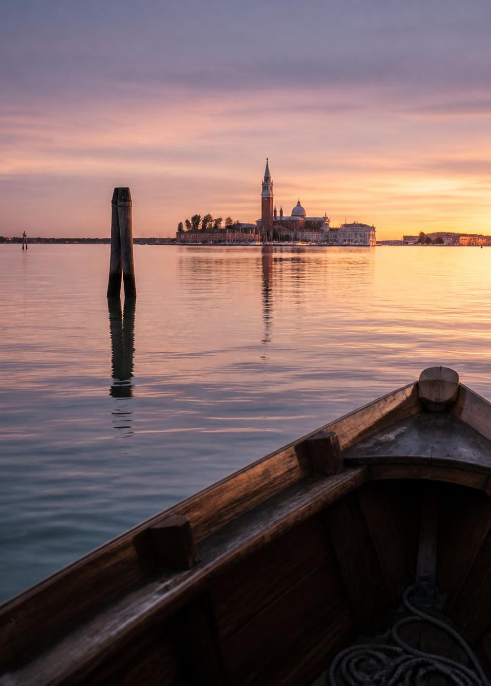 Venetian lagoon at dusk seen from a wooden boat with flat silver water and a distant bell tower