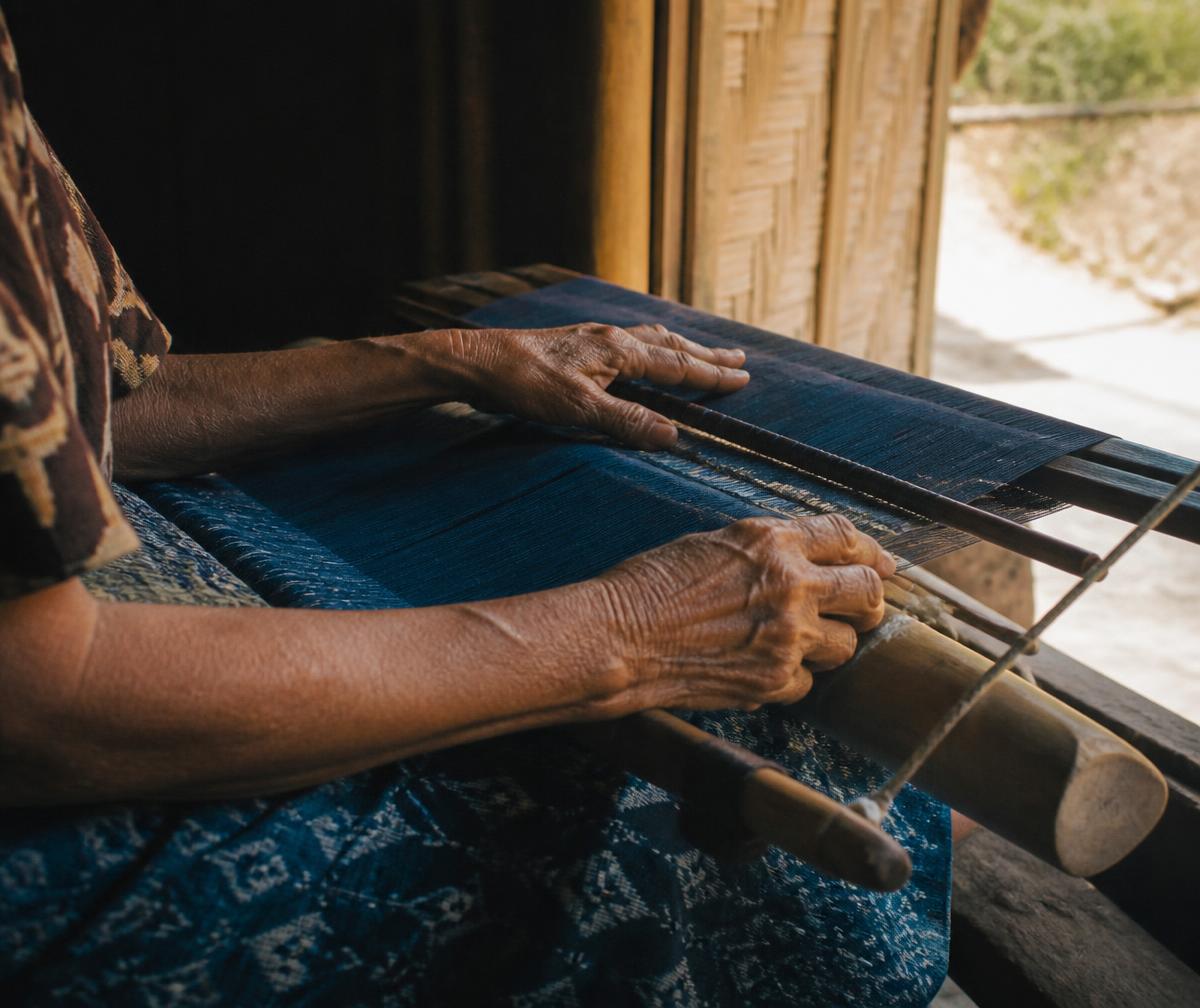 Hands of an elderly Sumbanese weaver working indigo cotton thread on a backstrap loom in warm side light