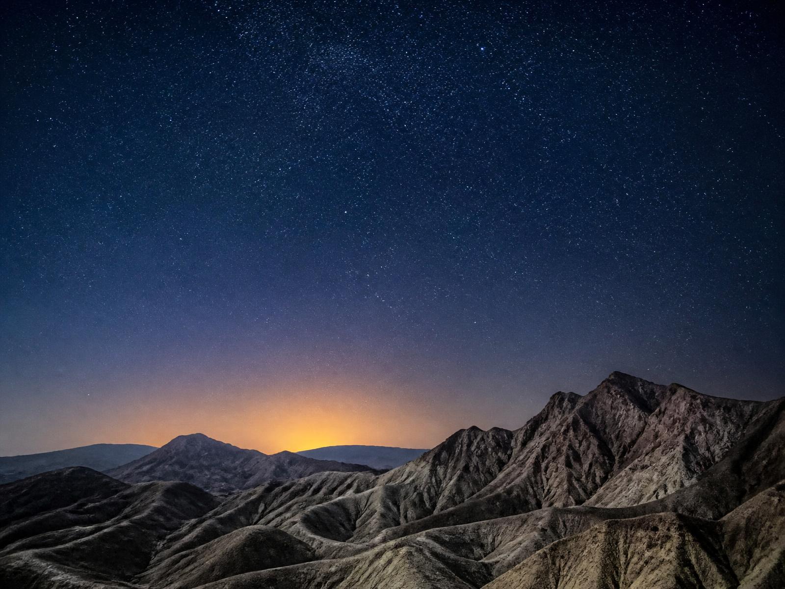 A vast star-filled desert night sky above bare rocky mountains with a faint warm glow on the horizon