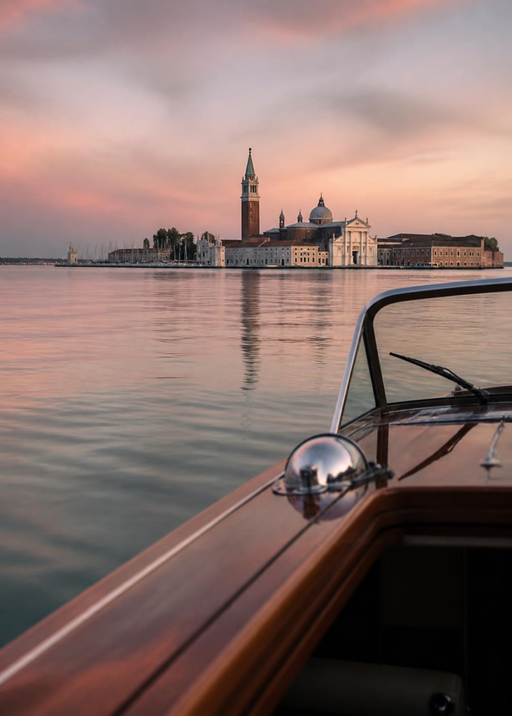 Venetian lagoon at dusk seen from a wooden boat, flat silver water and a distant bell tower