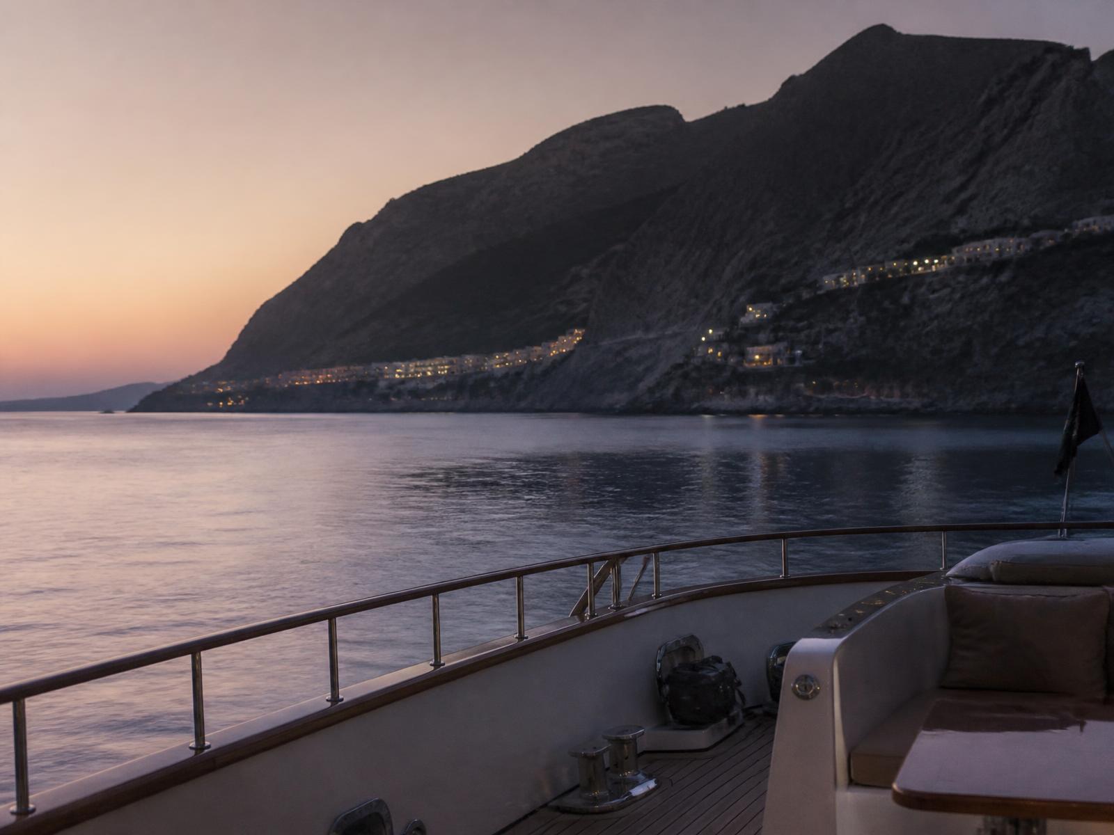 View from a yacht deck of a rugged Mediterranean island coastline at dusk with a calm pale sea and distant village lights
