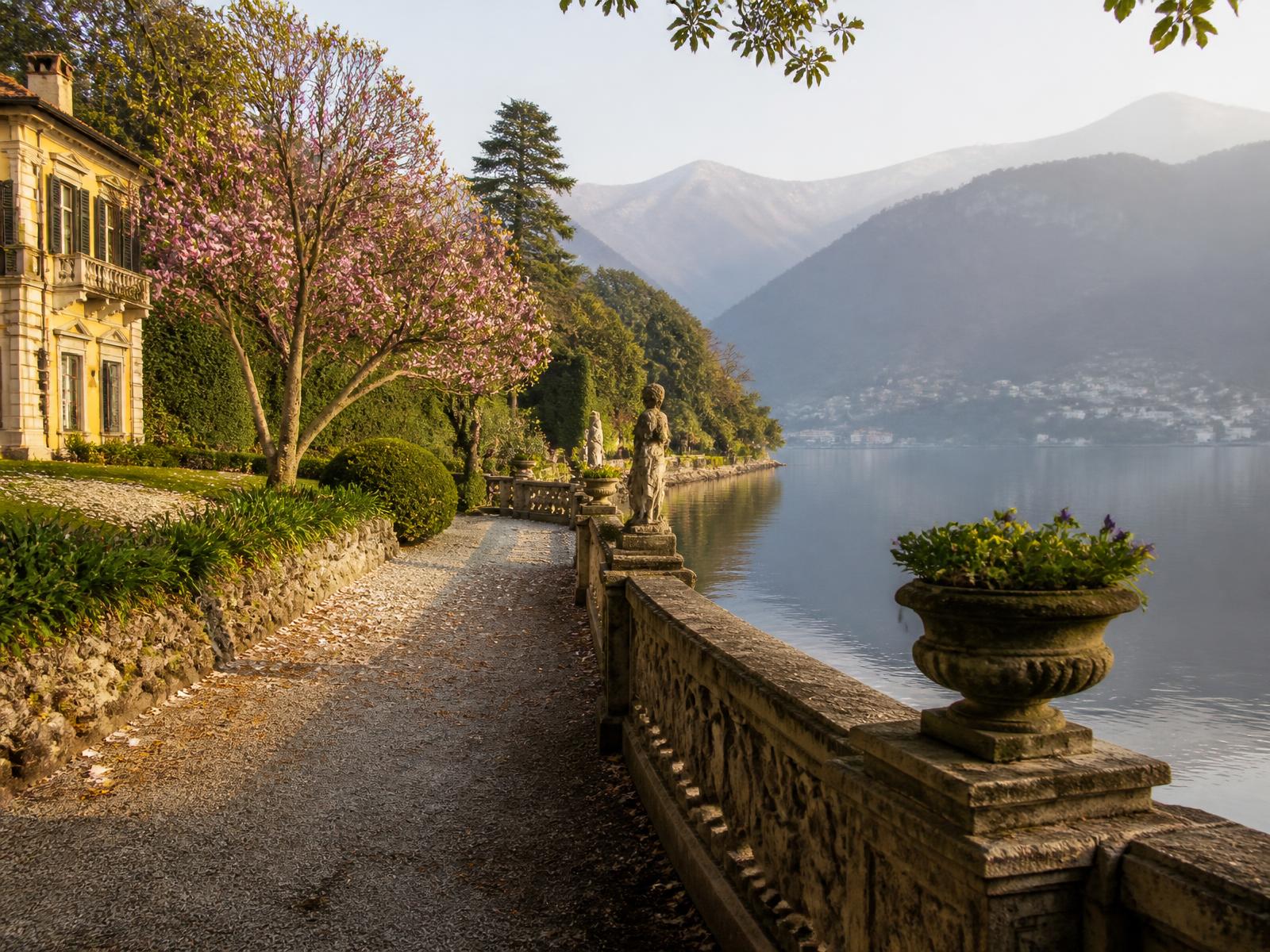 Historic Italian lakeside villa garden on Lake Como with magnolia trees, stone balustrade and mountains in morning haze