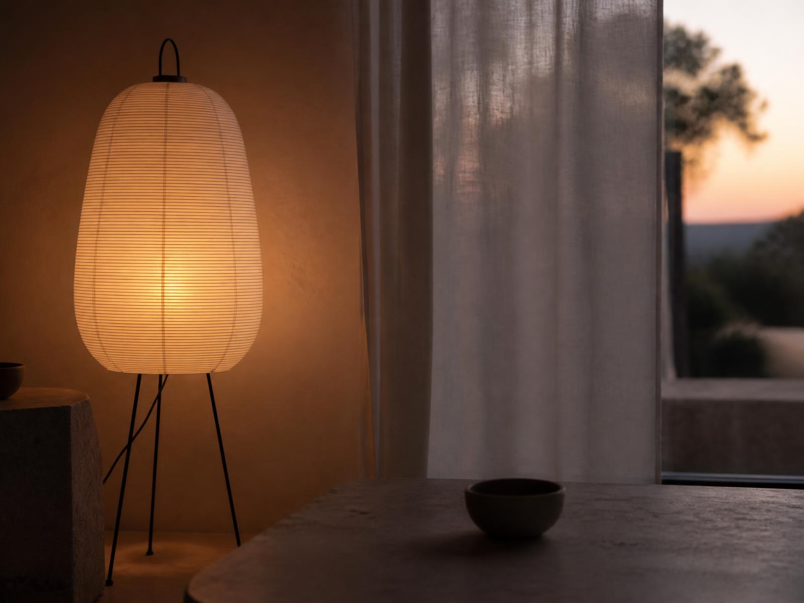 Warm leuchtende Papierschirm-Stehlampe neben einer Putzwand und einem Leinenvorhang in der Dämmerung