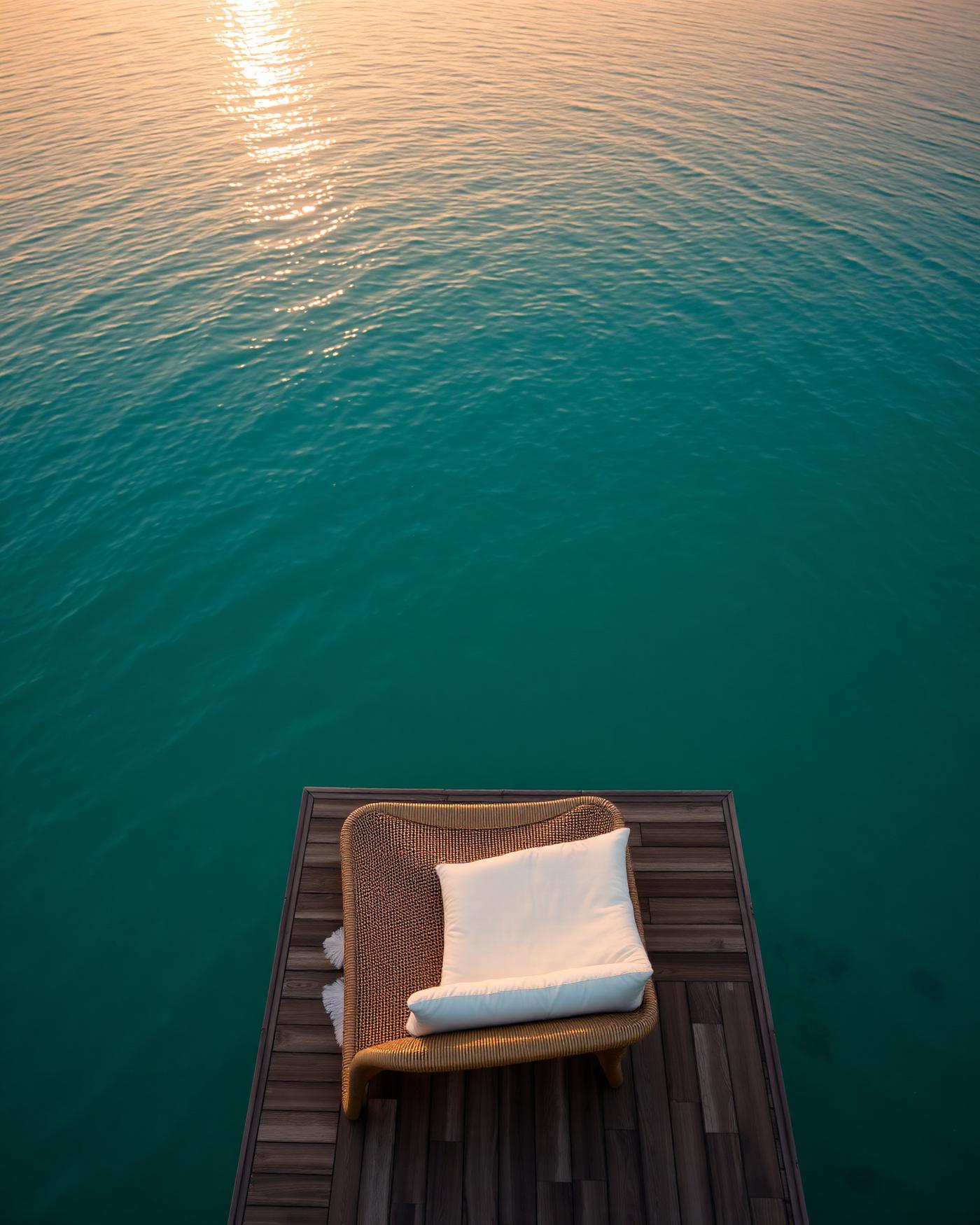 A teak deck with a woven lounger and linen cushion above still turquoise water at sunrise