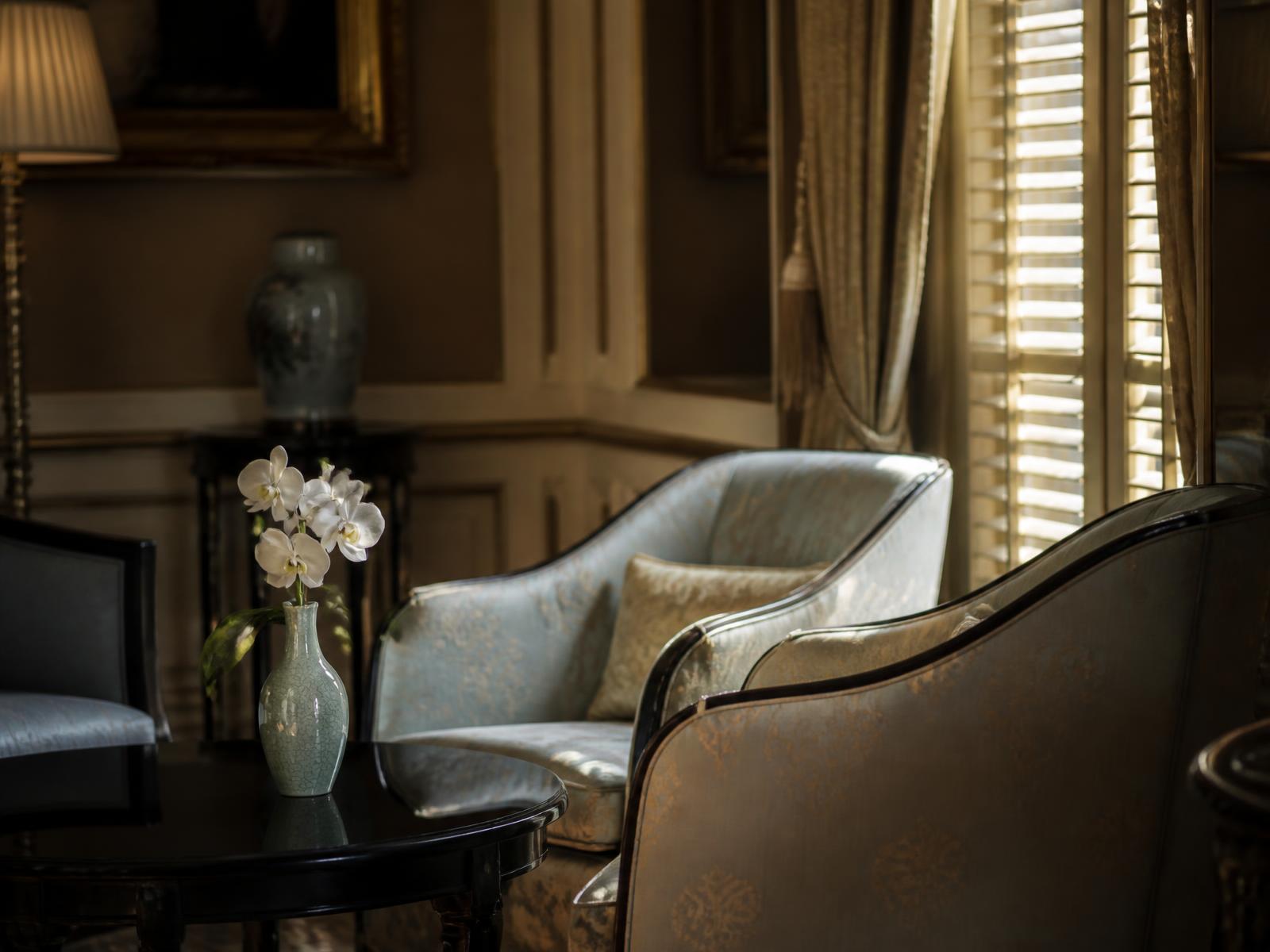 Silk-upholstered chairs and a single white orchid in a celadon vase beside a shuttered window