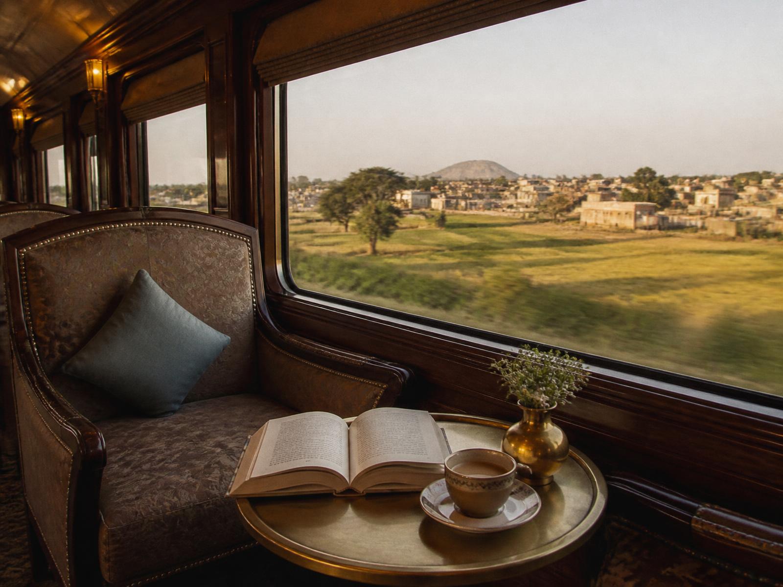 An observation lounge on an Indian luxury train with armchairs, a cup of masala chai and an open book on a brass table, rural India passing the window in golden light