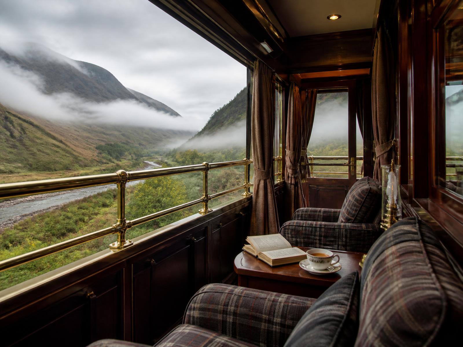 Der offene Aussichtswagen eines schottischen Luxuszugs mit einem Messinggeländer, Tartan-Sesseln, einem aufgeschlagenen Buch und einer Tasse Tee, ein nebliges Highland-Tal zieht vorbei