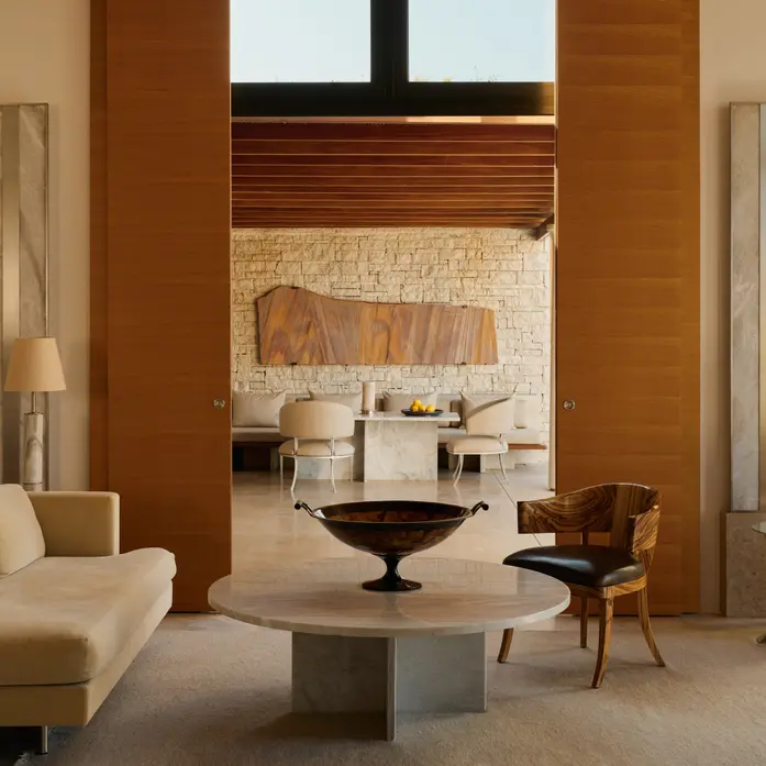 Wohnbereich eines Amanzoe-Pavillons mit hohen Holzpaneelen, heller Sitzlandschaft, rundem Marmortisch und Blick zum Essbereich vor Natursteinwand