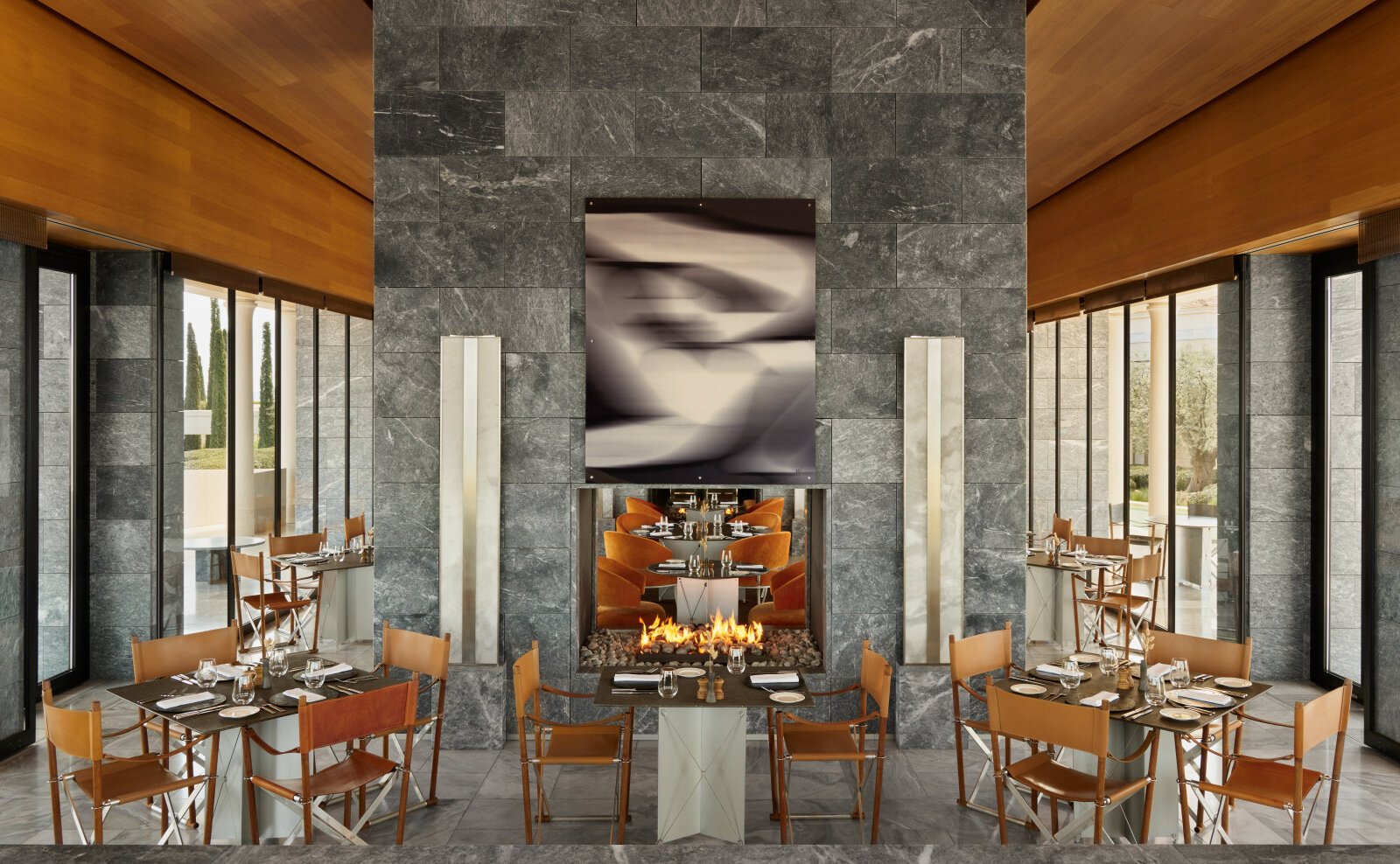 Hauptrestaurant in Amanzoe: Tische mit Regiestühlen vor einer grauen Steinwand mit offenem Feuer, seitlich raumhohe Fenster zu Zypressen