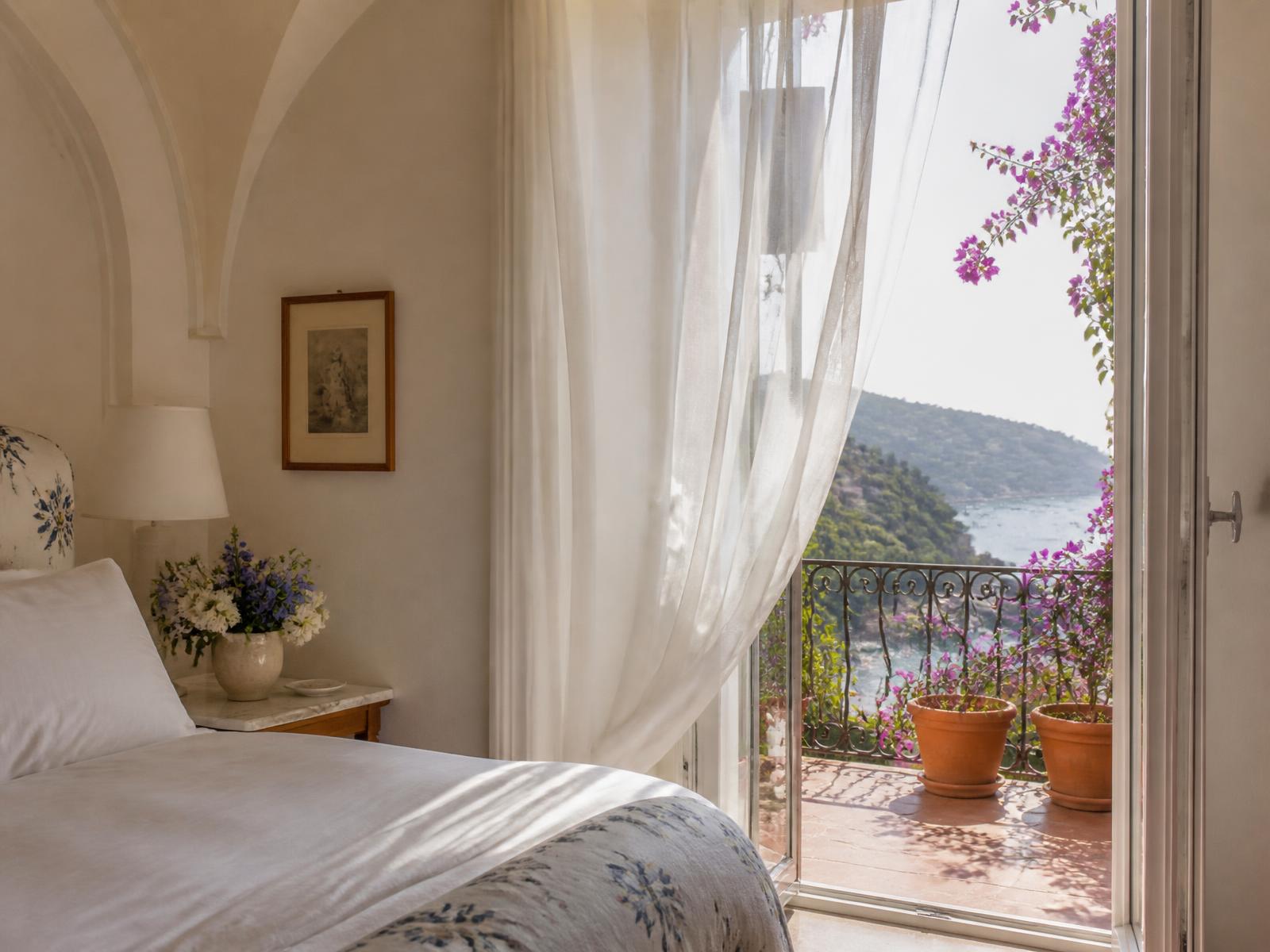 Riviera hotel bedroom with white linen, Italian printed fabrics and open doors onto a balcony with bougainvillea and the sea beyond