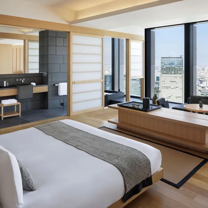 Zimmer mit hellem Holz, Shoji-Schiebewänden, niedrigem Bett und offenem Bad, dahinter ein raumhohes Fenster über Tokio